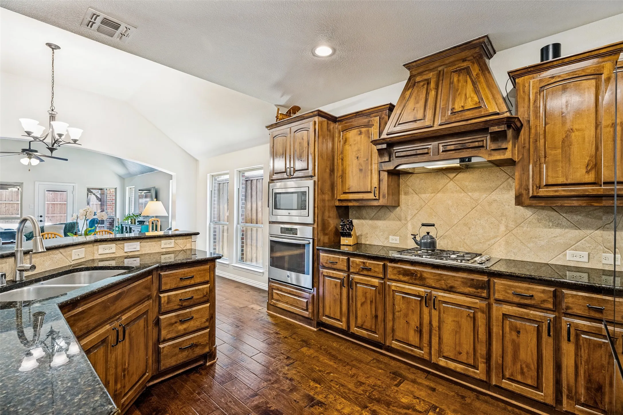 Vaulted ceiling kitchen featuring wood-finish cabinetry, dark countertops, a built-in oven, and a gas cooktop with an ornate range hood