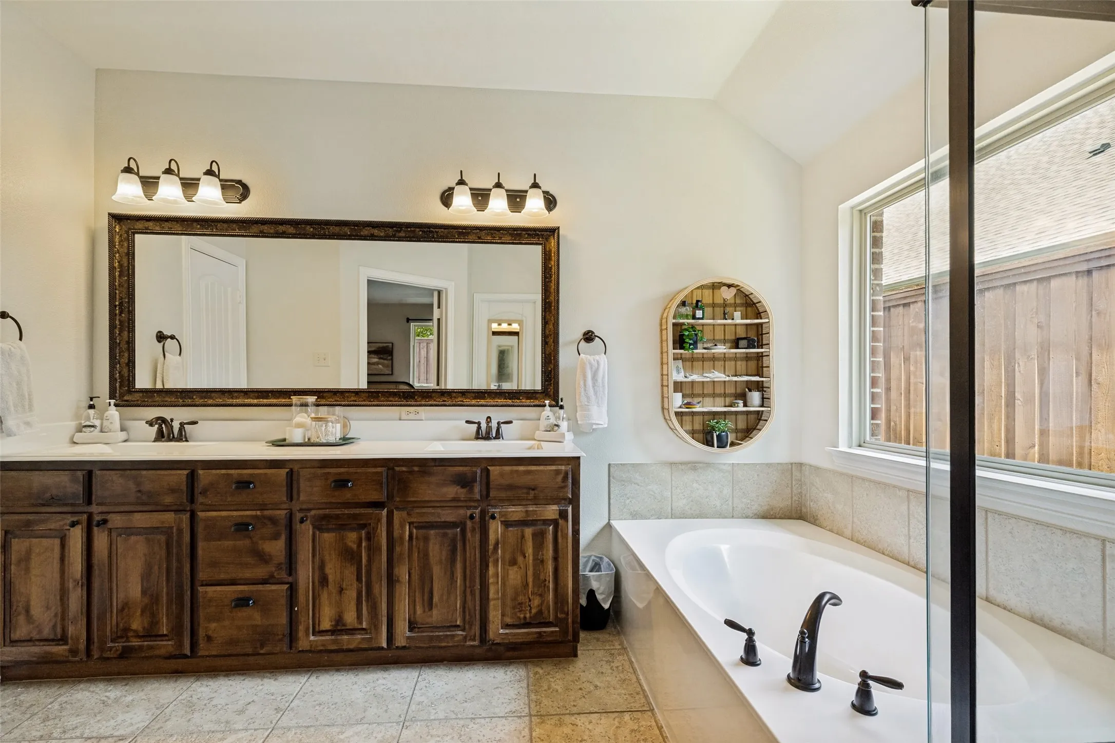 Dual vanity with dark wood cabinetry and white countertops, large framed mirror, recessed bathtub with dark fixtures, separate glass-enclosed shower, and a large window