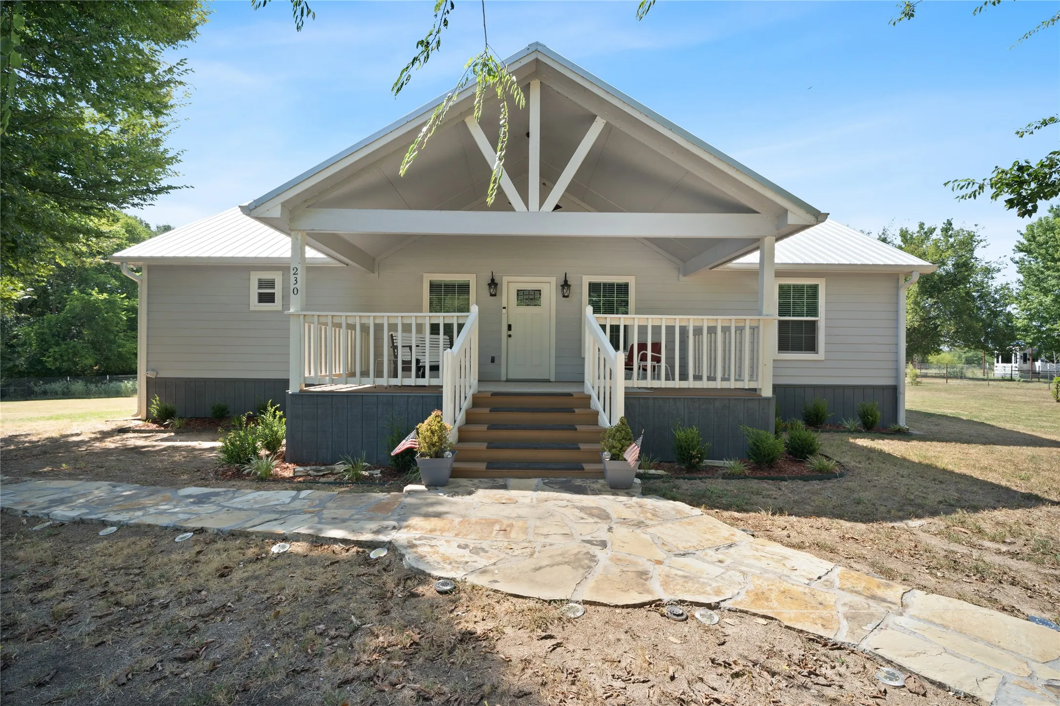 Gabled front porch with exposed beam detailing, light gray siding, white metal roof, wood-look steps, and a flagstone walkway