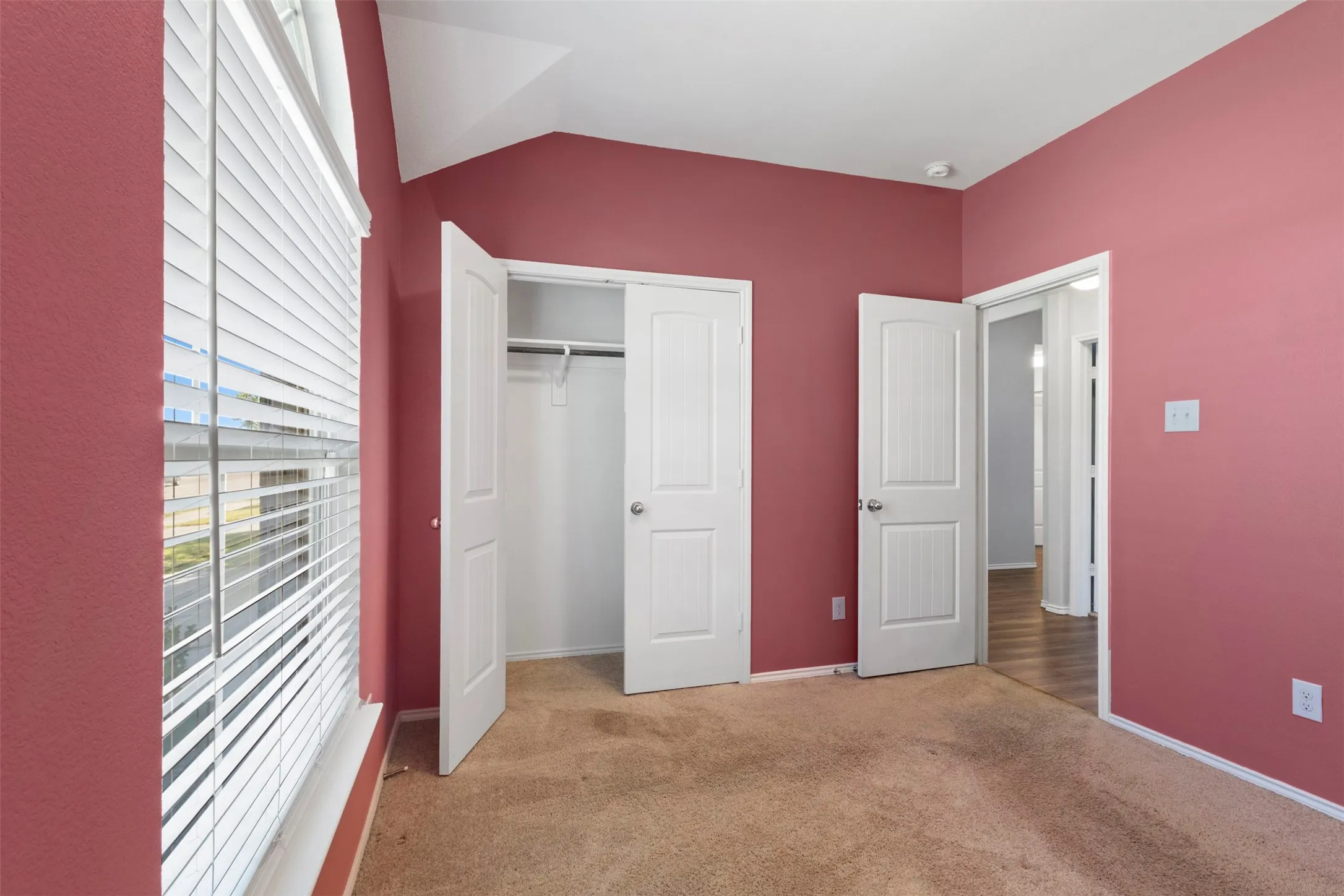 Angled ceiling with white trim, built-in closet with bifold doors, window with horizontal blinds, and carpeting