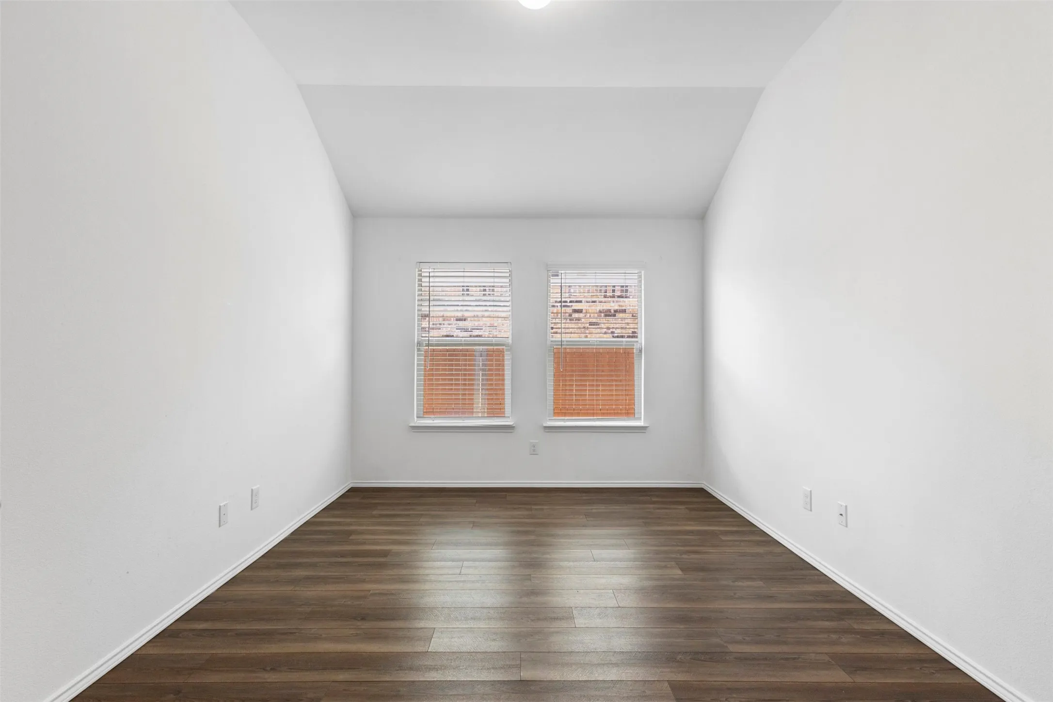 Vaulted ceiling with a flush-mount light fixture, wood-finish flooring, and two windows with blinds