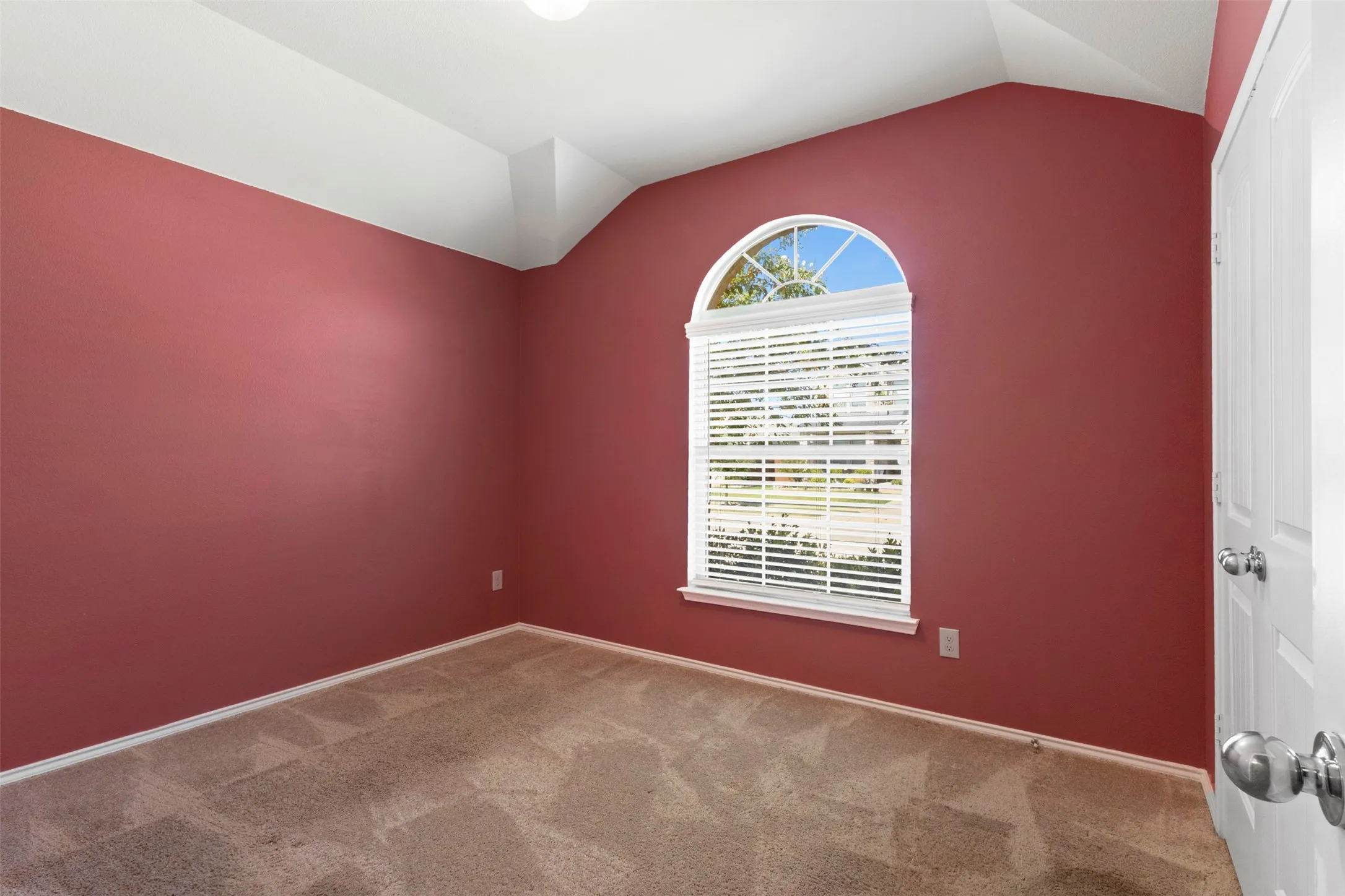 Arched window with blinds, vaulted ceiling, neutral carpet flooring, and a white paneled door