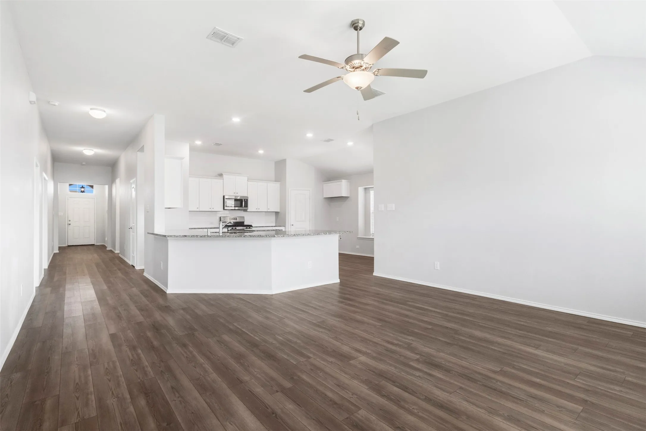 Open-concept living and kitchen area featuring gleaming wood-finish flooring throughout, recessed ceiling lighting, a ceiling fan with integrated light fixture, and a curved white cabinetry island with quartz countertop and stainless steel appliances