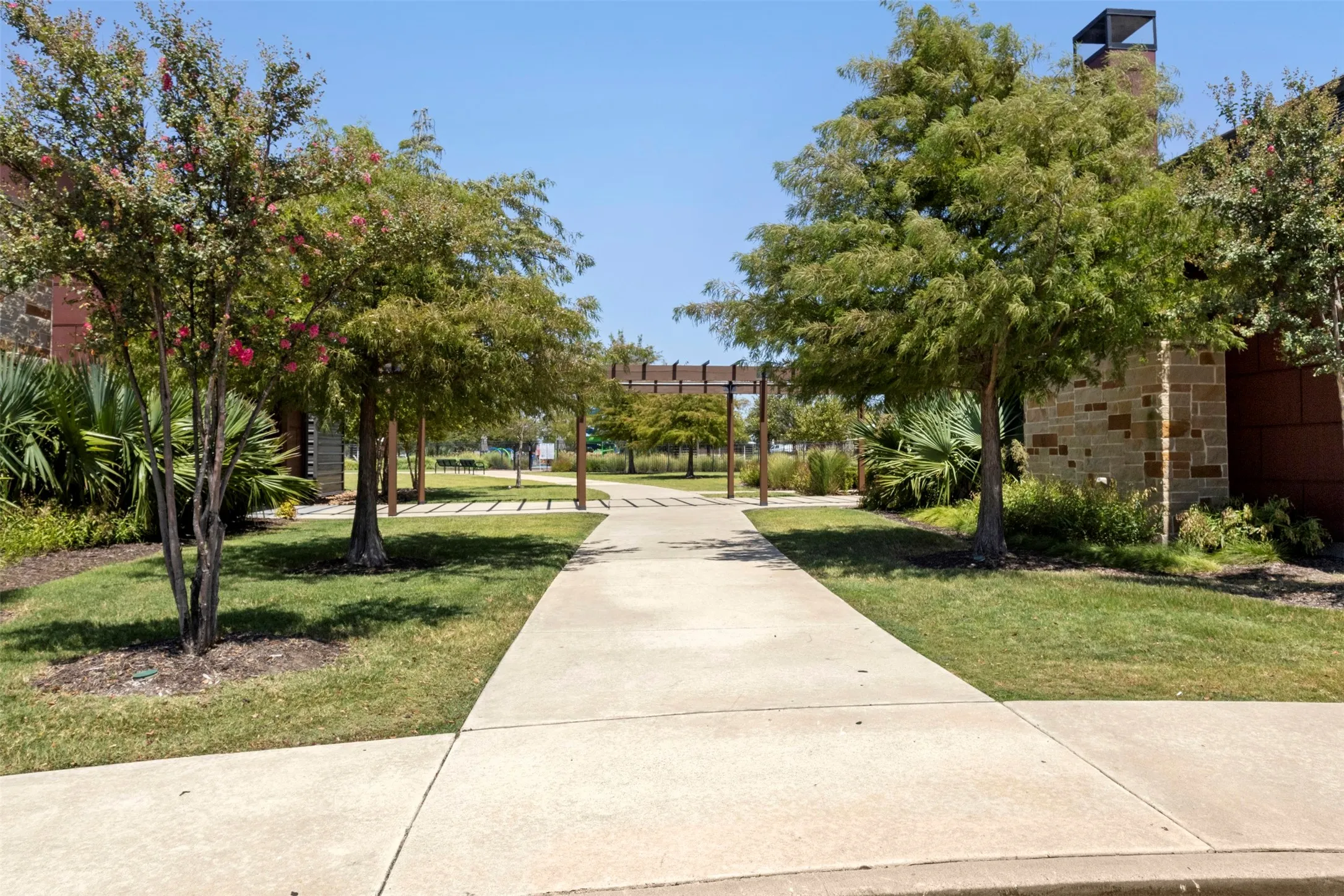 Concrete pathway leading to a landscaped green area with mature trees and a stone-clad structure