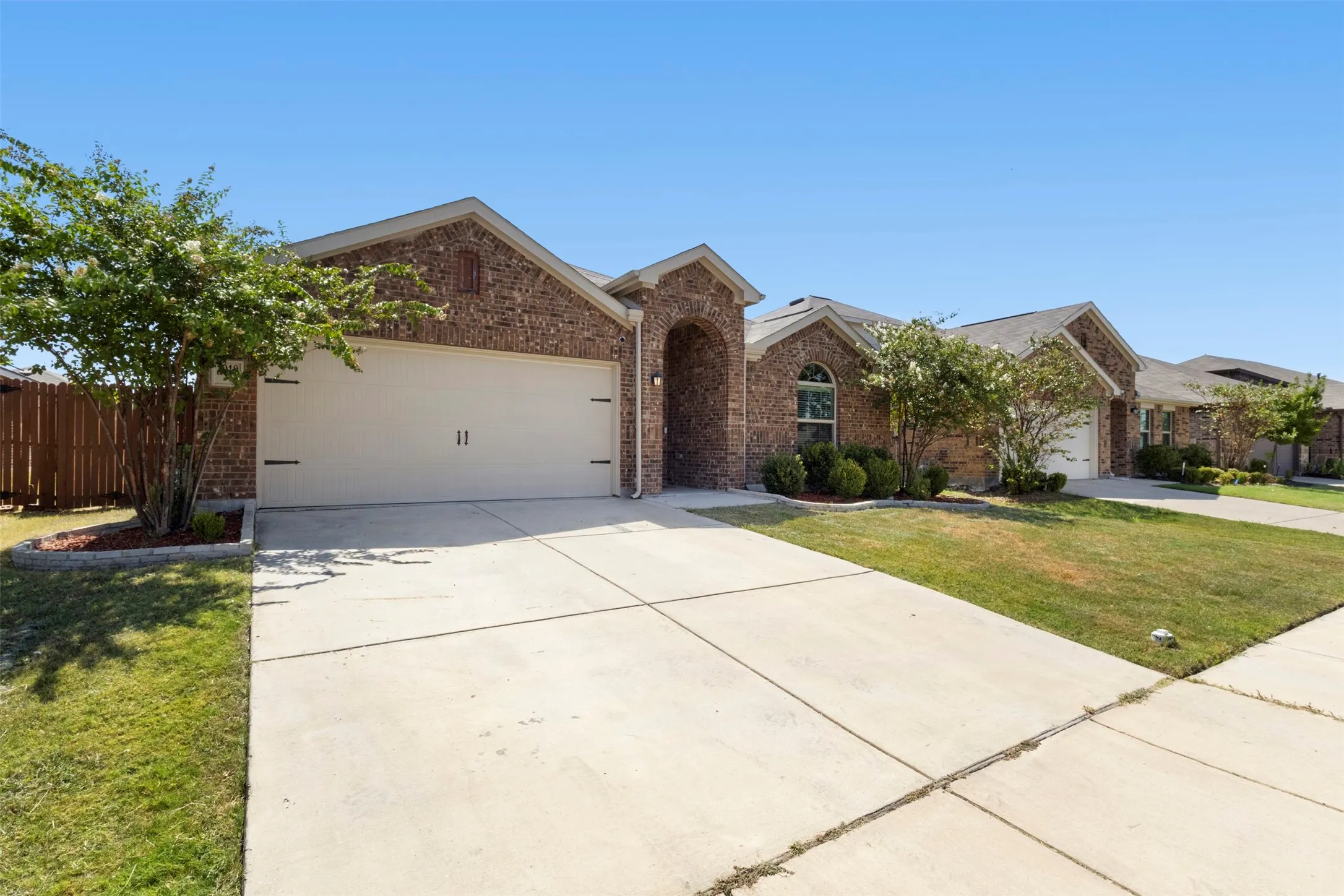 Brick exterior with arched entry, attached garage, concrete driveway, and established front landscaping