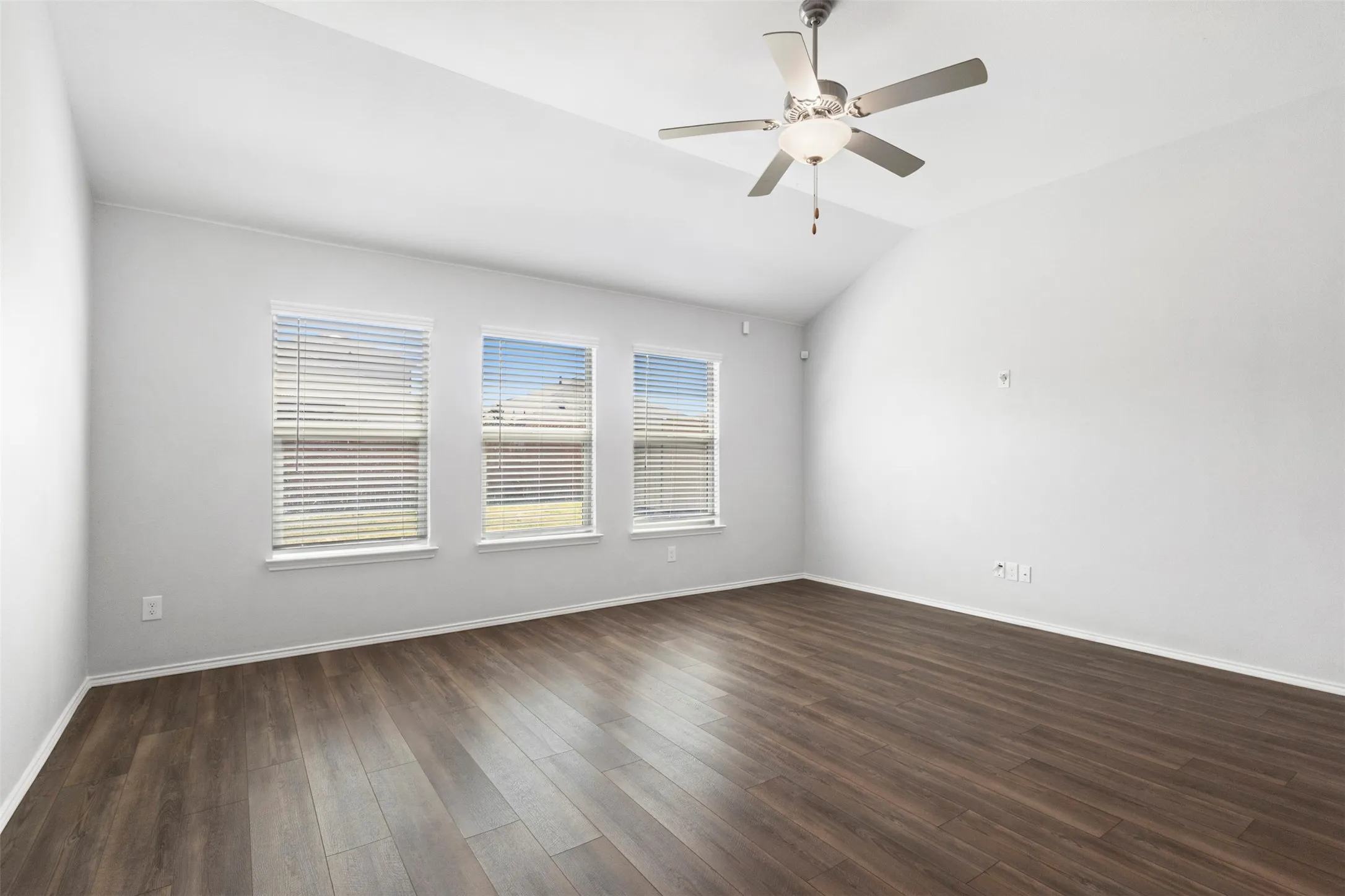 Vaulted ceiling, wood-finish flooring, three windows with blinds, and a ceiling fan with light fixture