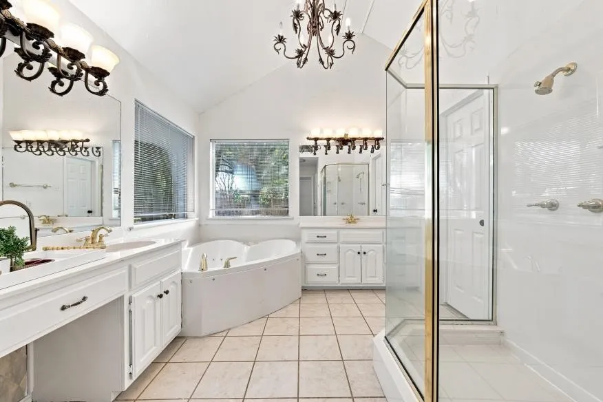Dual vanity with gold-finish fixtures and white cabinetry, separate glass-enclosed shower, jetted soaking tub, tiled flooring, and vaulted ceiling