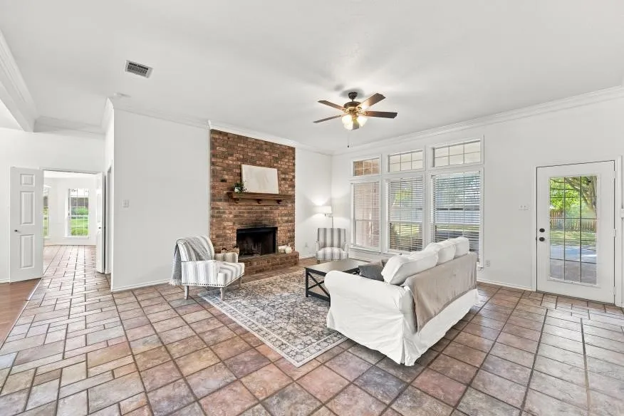 Exposed brick fireplace with a wood mantle, ceiling fan, tile flooring, glass-paned exterior door, and multiple windows