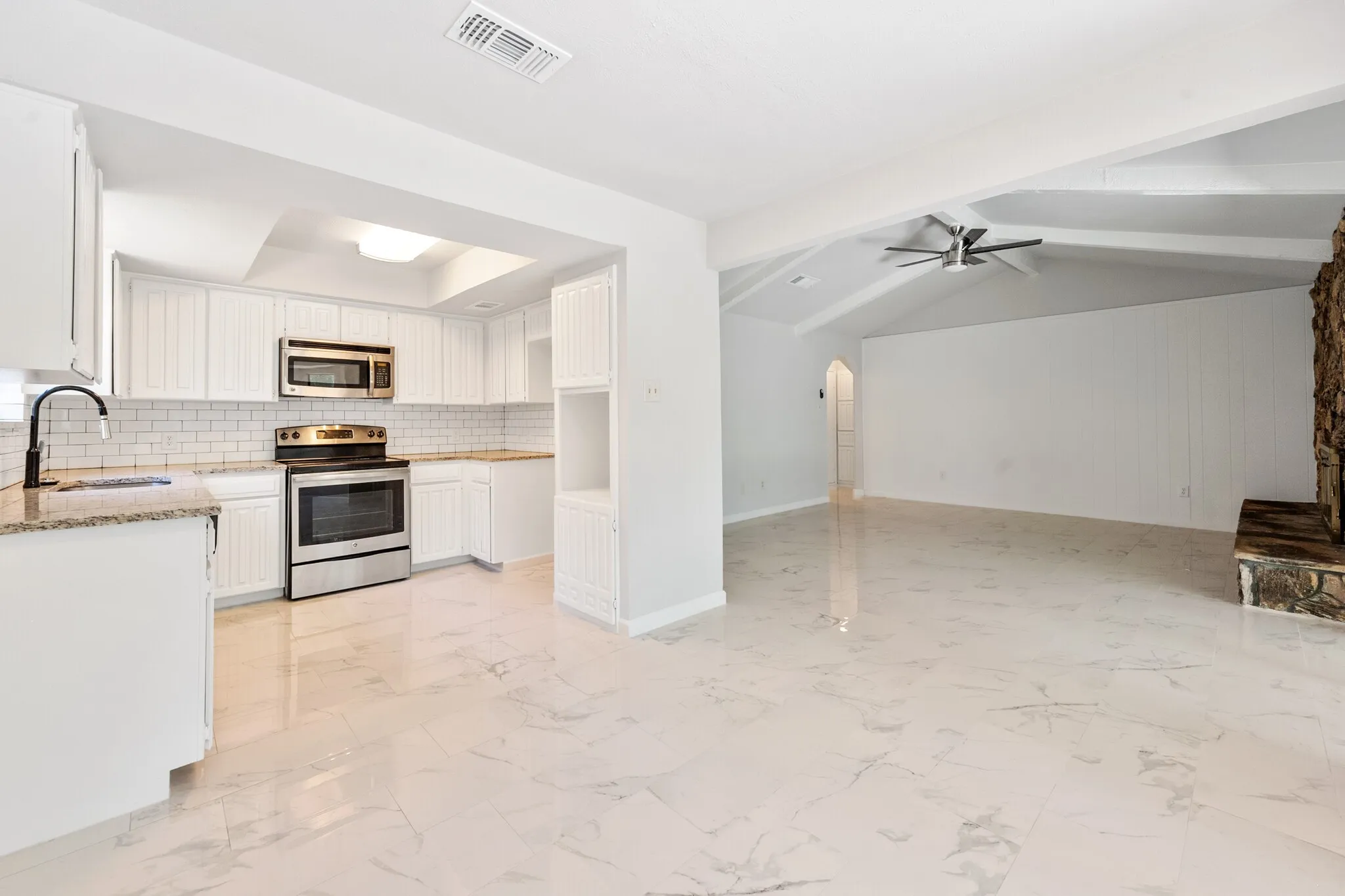 Open-concept living space featuring white cabinetry, stainless steel appliances, white subway tile backsplash, granite-style countertops, and a stone-clad fireplace