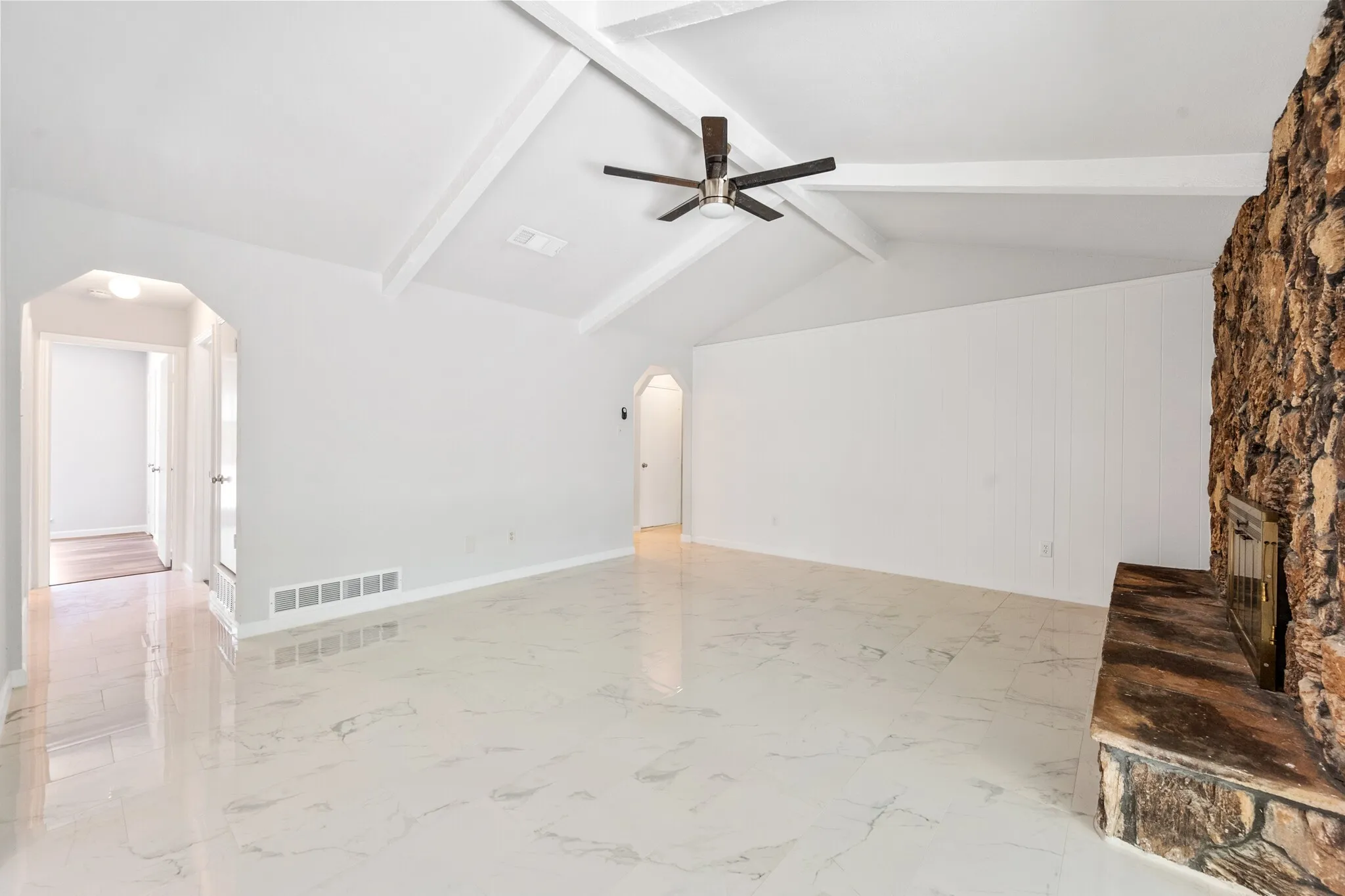 Vaulted ceiling with exposed beams, wood-look ceiling fan, stone-clad fireplace, and large format tile flooring