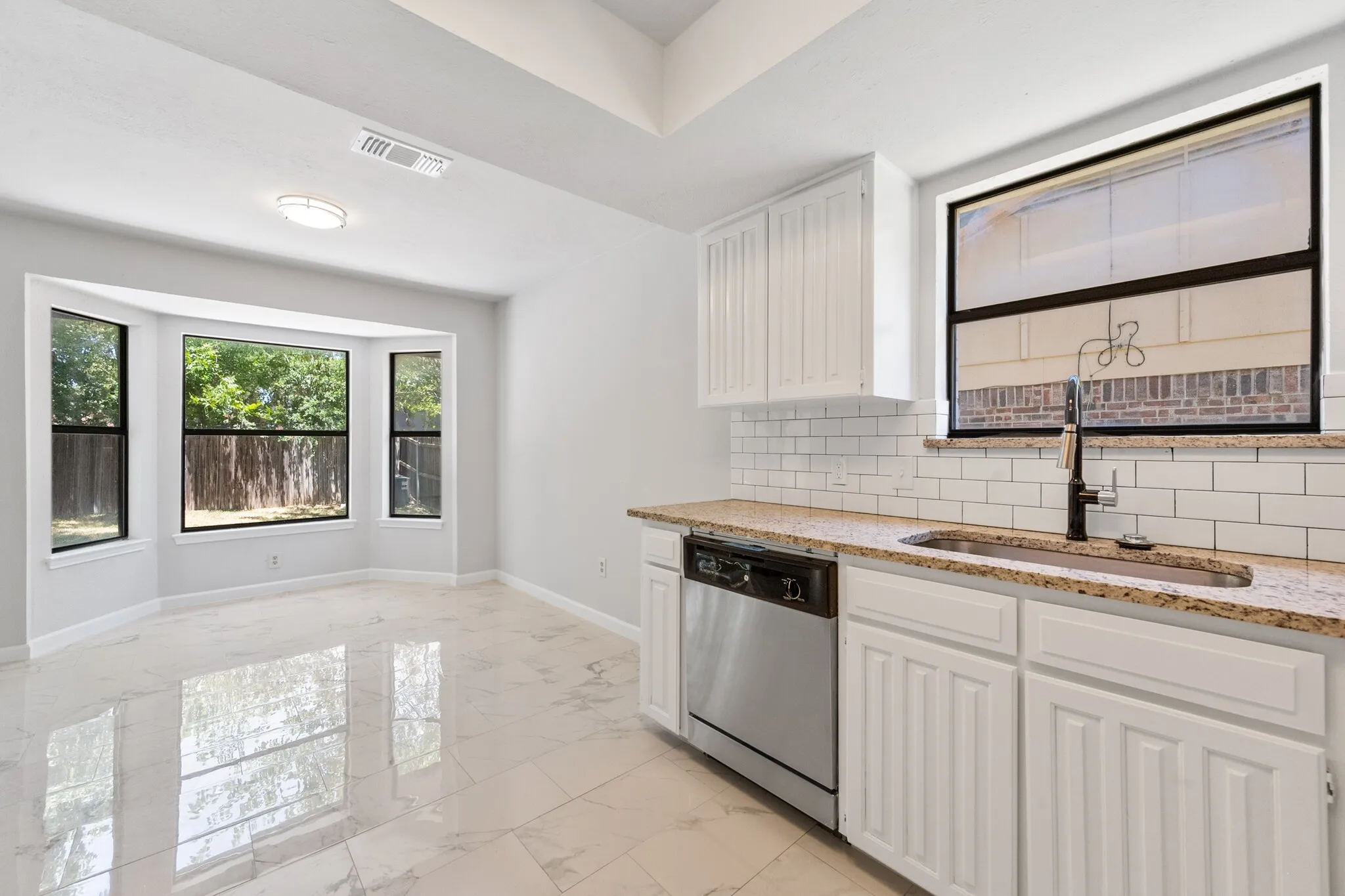 White shaker cabinetry, stone-finish countertops, stainless steel dishwasher, white subway tile backsplash, and a bay window