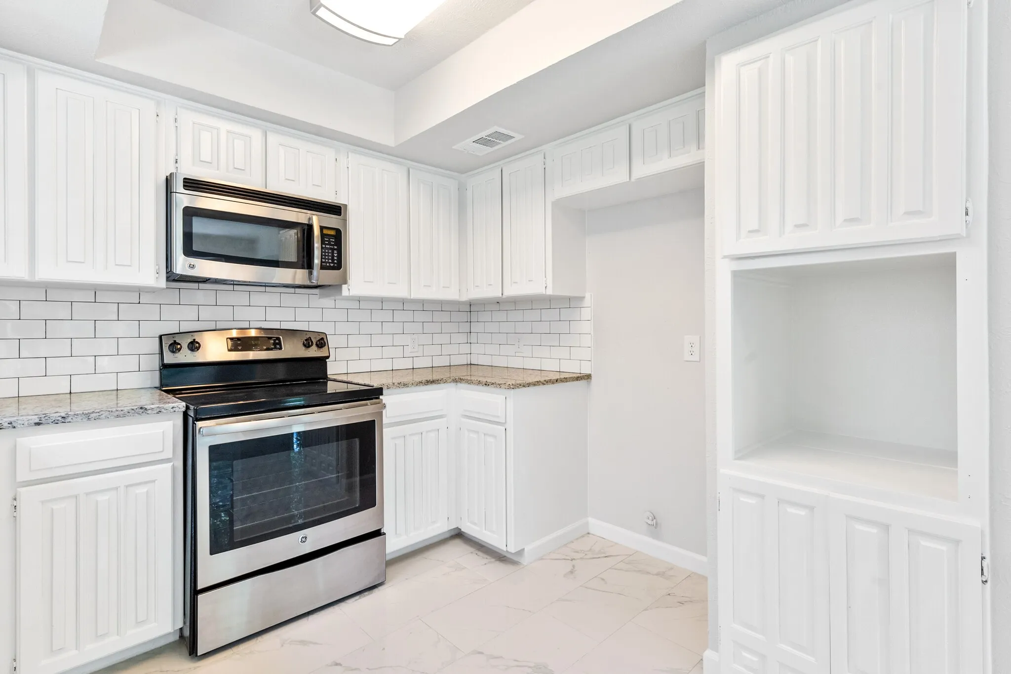 White shaker-style cabinetry, stainless steel oven with an electric cooktop, stainless steel microwave, light-toned subway tile backsplash, and stone countertops