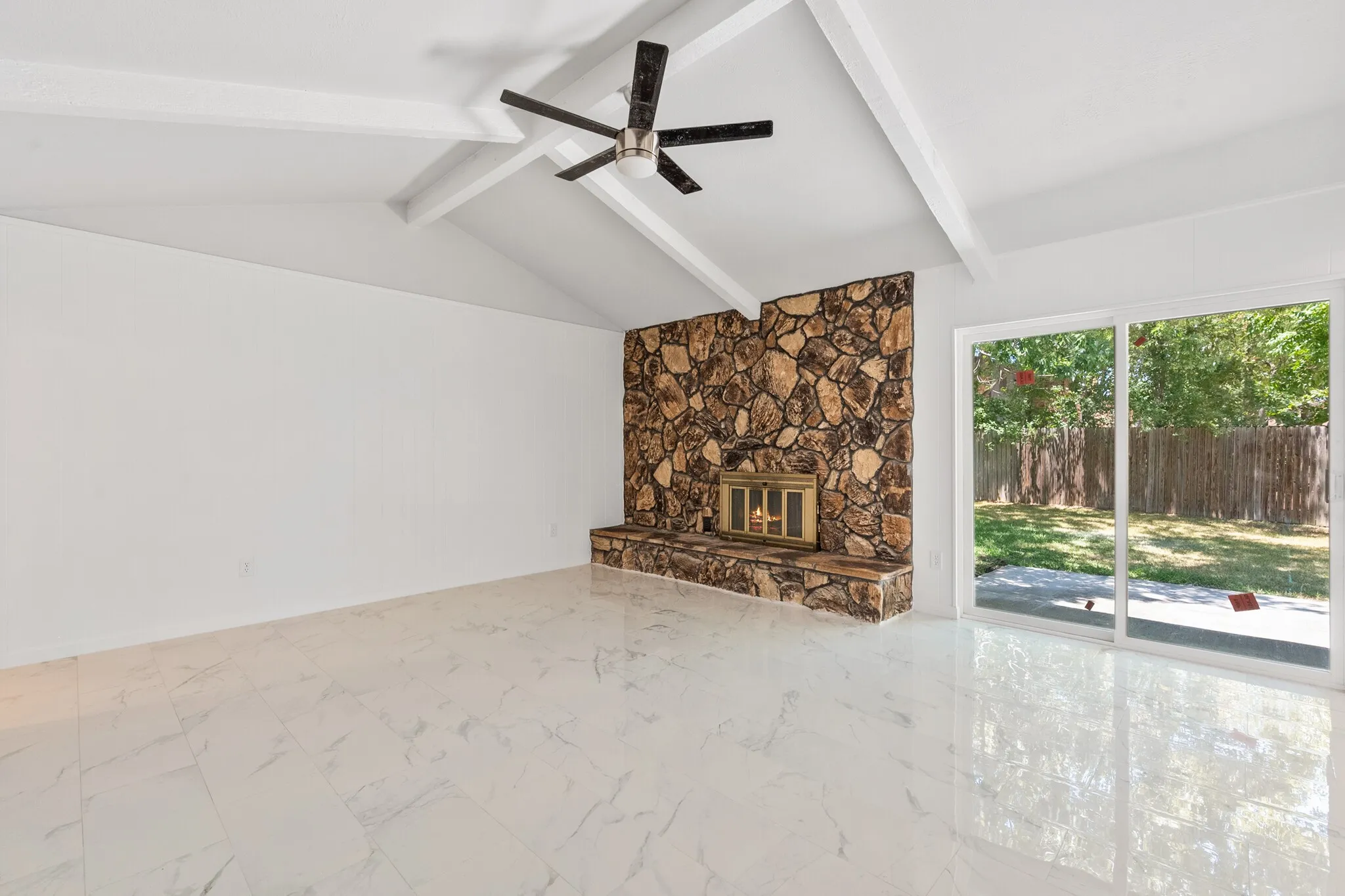 Vaulted ceiling with exposed beams, ceiling fan, natural stone fireplace, large sliding glass doors, and light-colored tile flooring