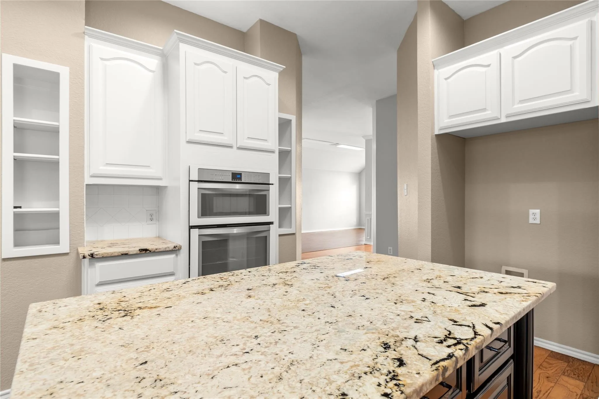 Kitchen island with a light-toned natural stone countertop, white upper cabinetry, stainless steel double ovens, and open shelving
