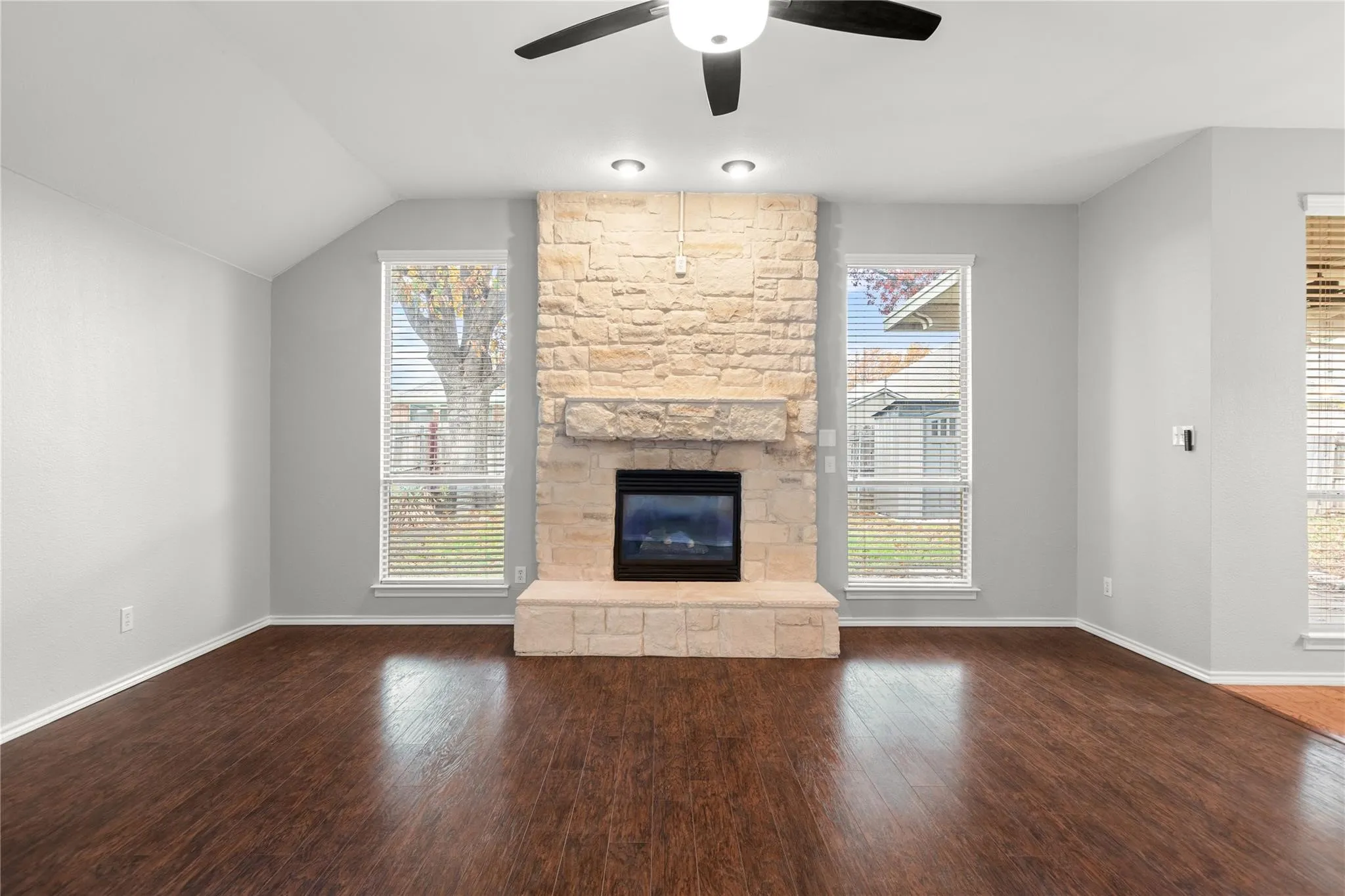 Stone-clad fireplace with hearth, wood-finish flooring, vaulted ceiling, and ceiling fan with integrated lighting