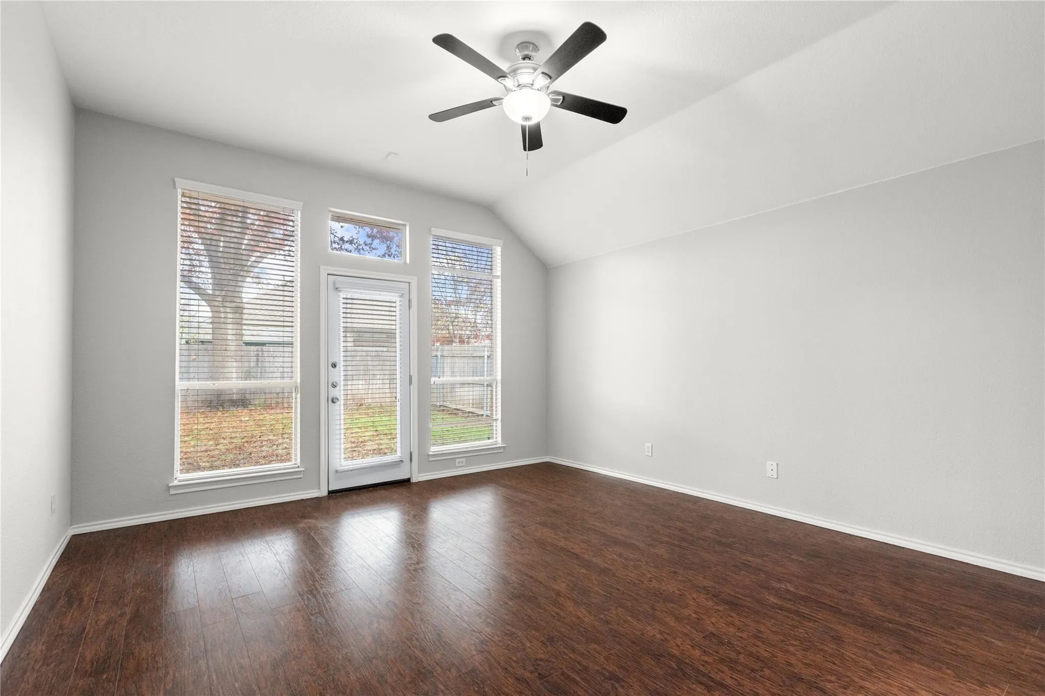 Vaulted ceiling, wood-finish flooring, ceiling fan, multiple windows with blinds, and a glass-paneled door