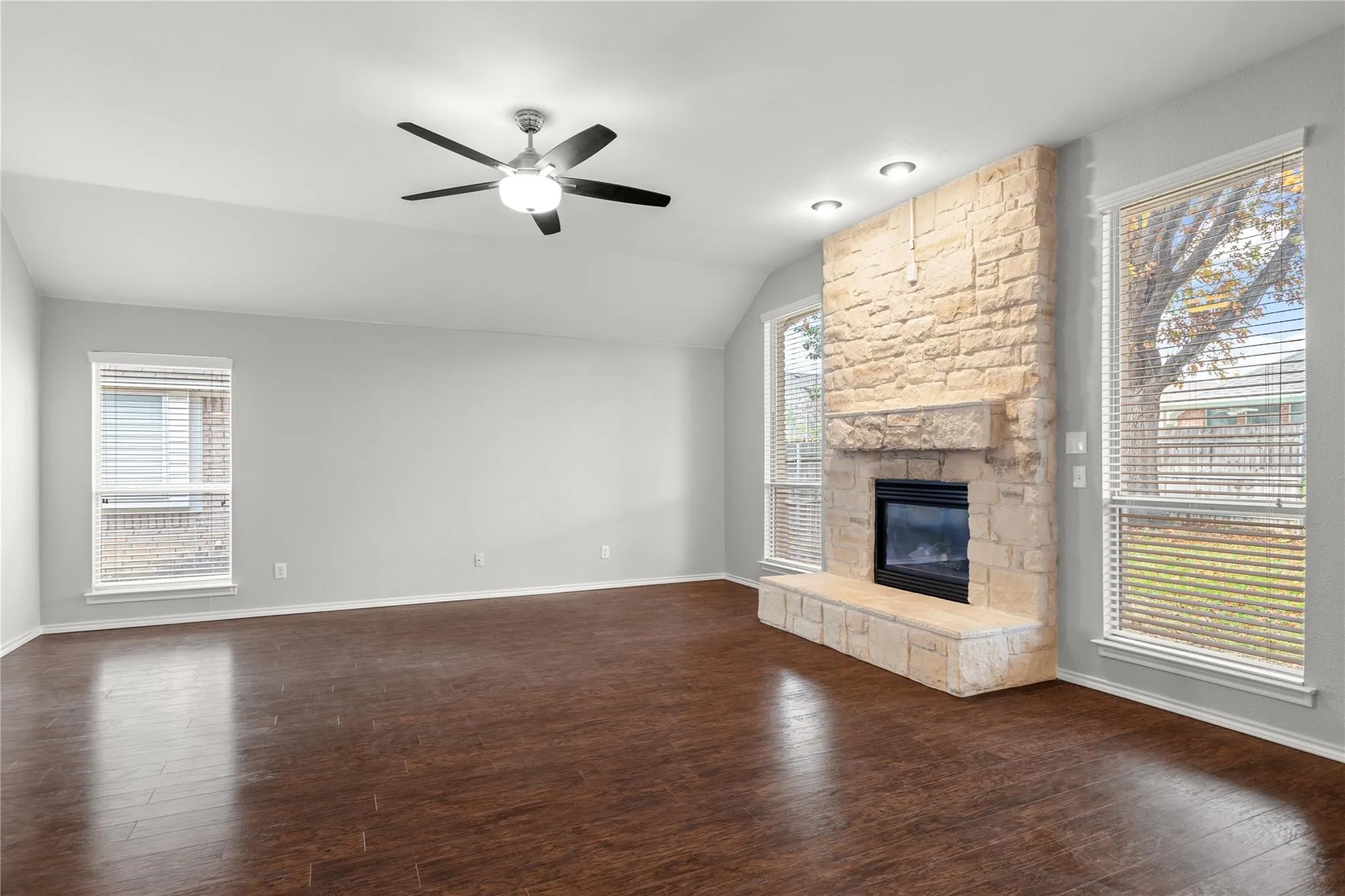 Vaulted ceiling with a ceiling fan, stone-clad fireplace with hearth, wood-finish flooring, and multiple windows