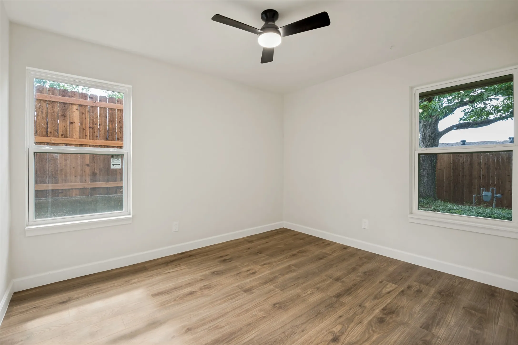 Wood-finish flooring, white walls, two windows with white trim, and a black ceiling fan with an integrated light