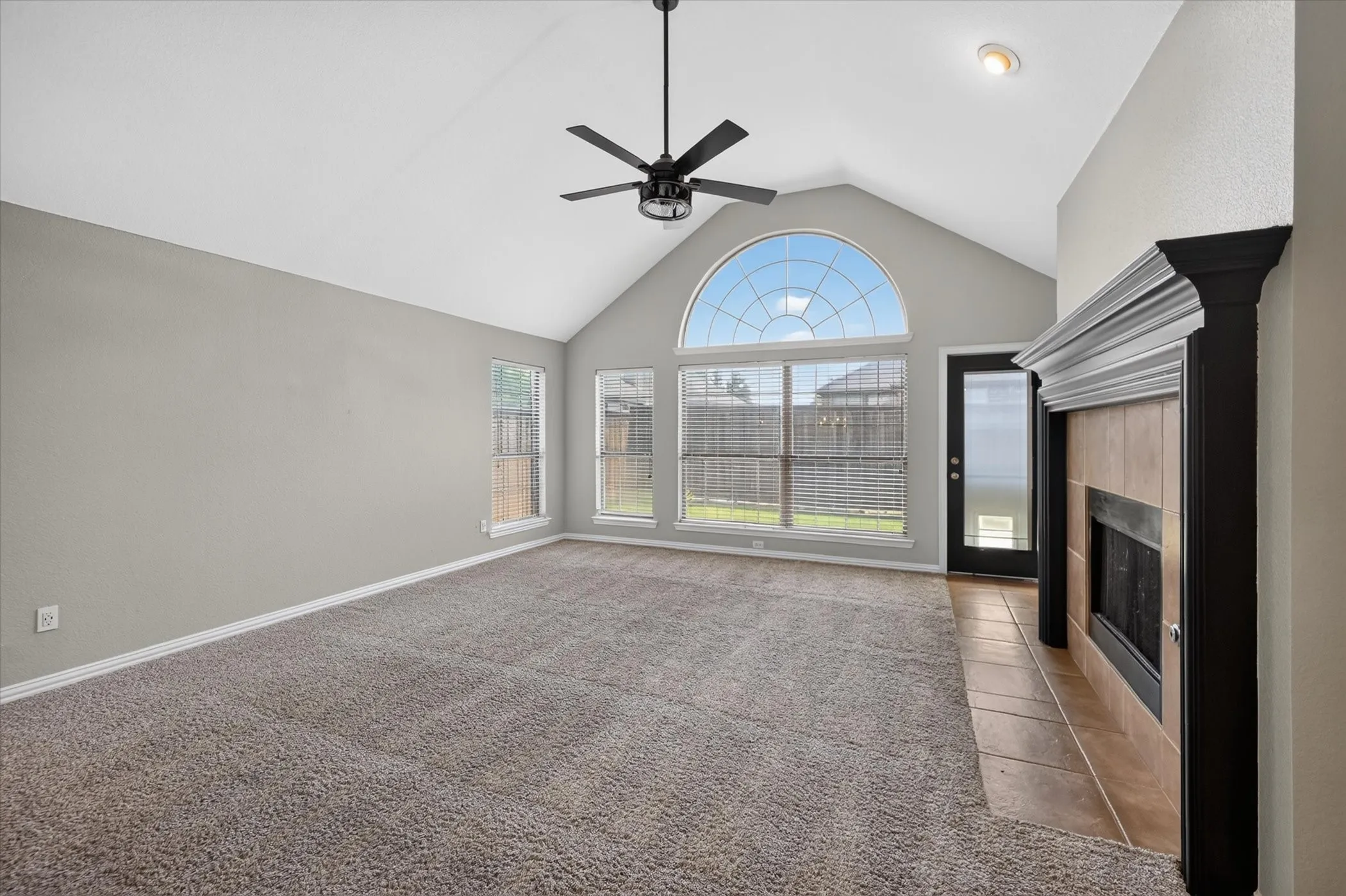 Vaulted ceiling with an installed ceiling fan, an arched transom window, a tiled fireplace with a dark wood mantel, and light gray wall paint