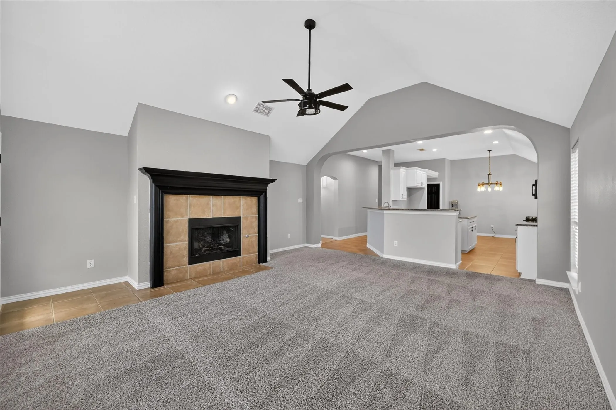 Vaulted ceilings, carpet flooring, a fireplace with a black mantel and tile surround, and an arched entryway to the kitchen with wood-finish flooring