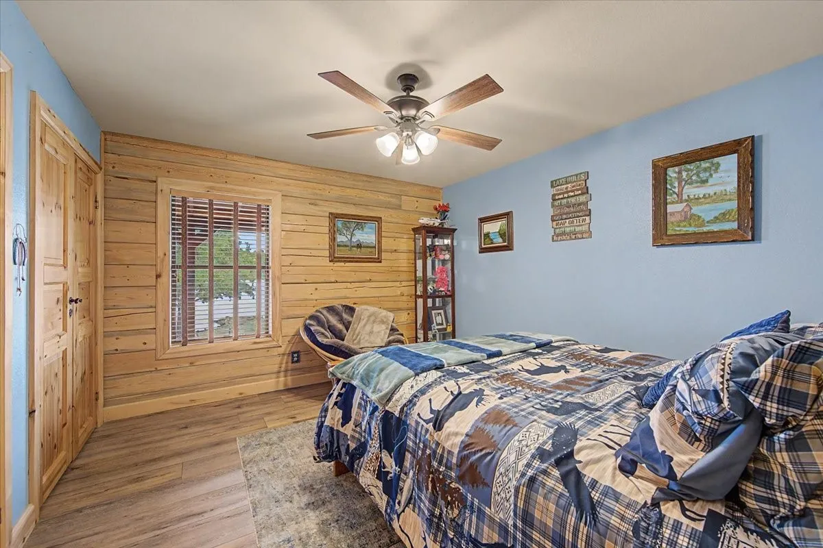 Wood-finish flooring, paneled wood wall, window with blinds, and ceiling fan