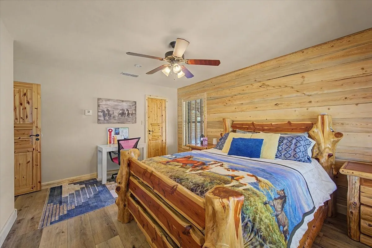 Wood-finish flooring, accent wall featuring horizontal wood paneling, wood-frame window with blinds, wood-paneled doors, and a ceiling fan with integrated lighting