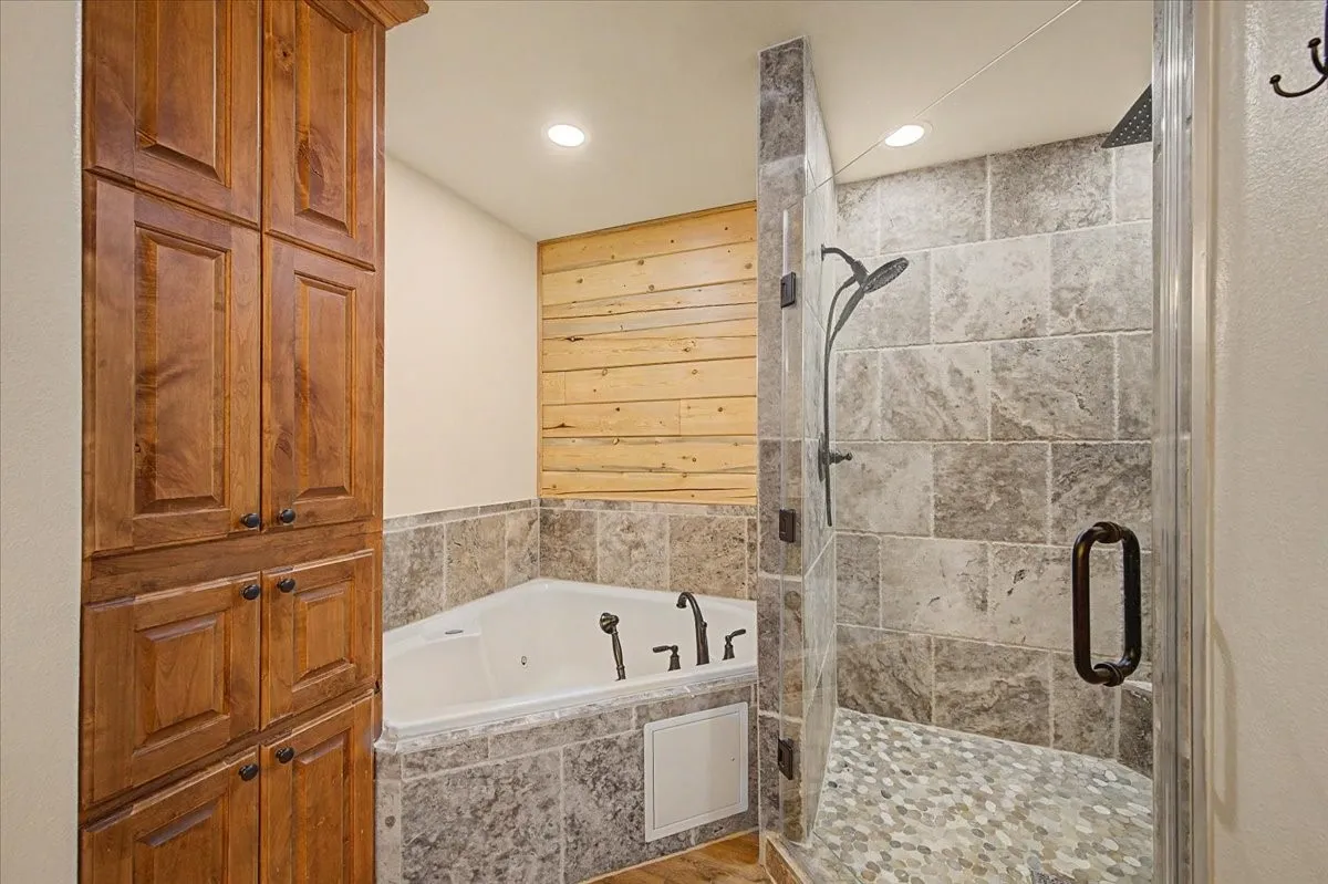 Built-in wood cabinetry, corner jetted tub, tiled tub surround, walk-in shower with glass enclosure, and pebble-tile shower floor