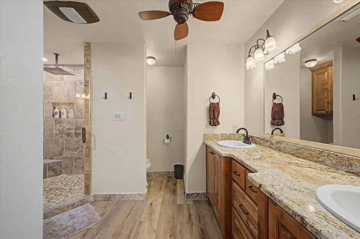 Wood-finish flooring, dual vanity with stone countertops, and a glass-enclosed shower with a built-in bench and rainfall showerhead