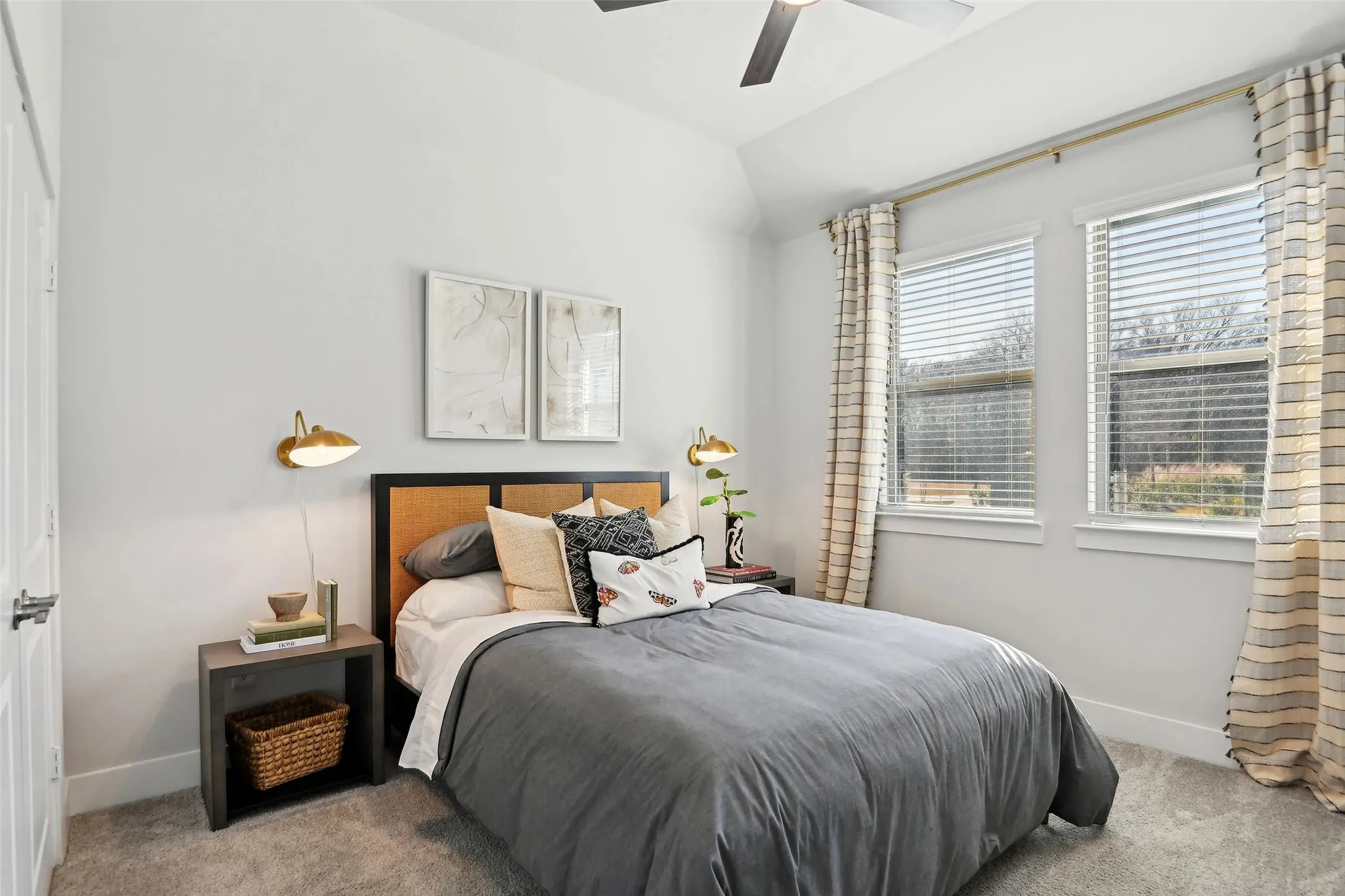 Vaulted ceiling with ceiling fan, two windows with blinds, wall-mounted light fixtures, wood-finish nightstand, and carpet flooring
