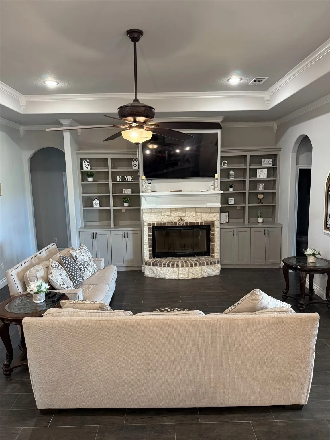 Built-in cabinetry with open shelving and lower storage, stone-clad fireplace with brick hearth, dark tile flooring, ceiling fan with integrated lighting, and crown molding