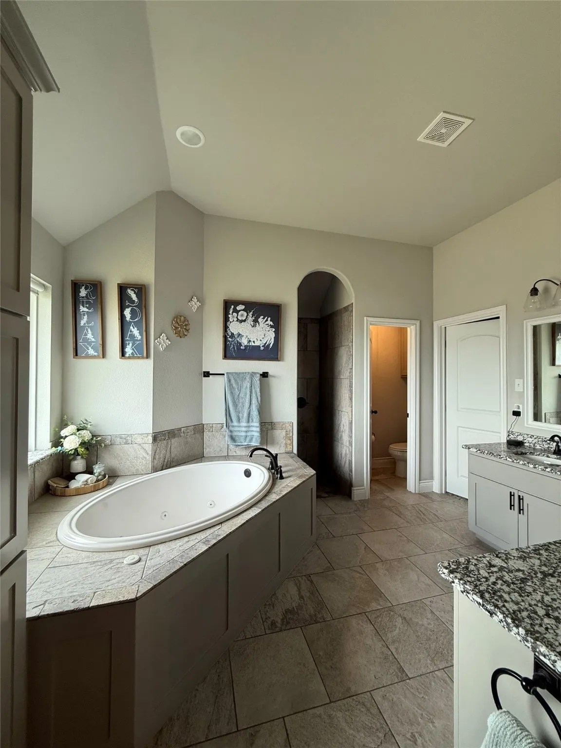 Soaking tub with tiled surround, dual vanity with granite-finish countertop, tiled flooring, and arched shower entry