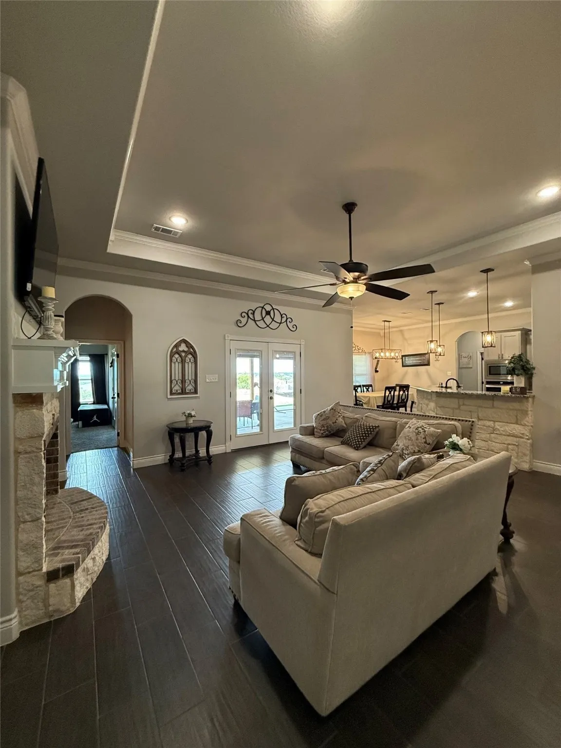Tray ceiling with recessed lighting, dark wood-finish flooring, stone-clad fireplace, French doors, and ceiling fan