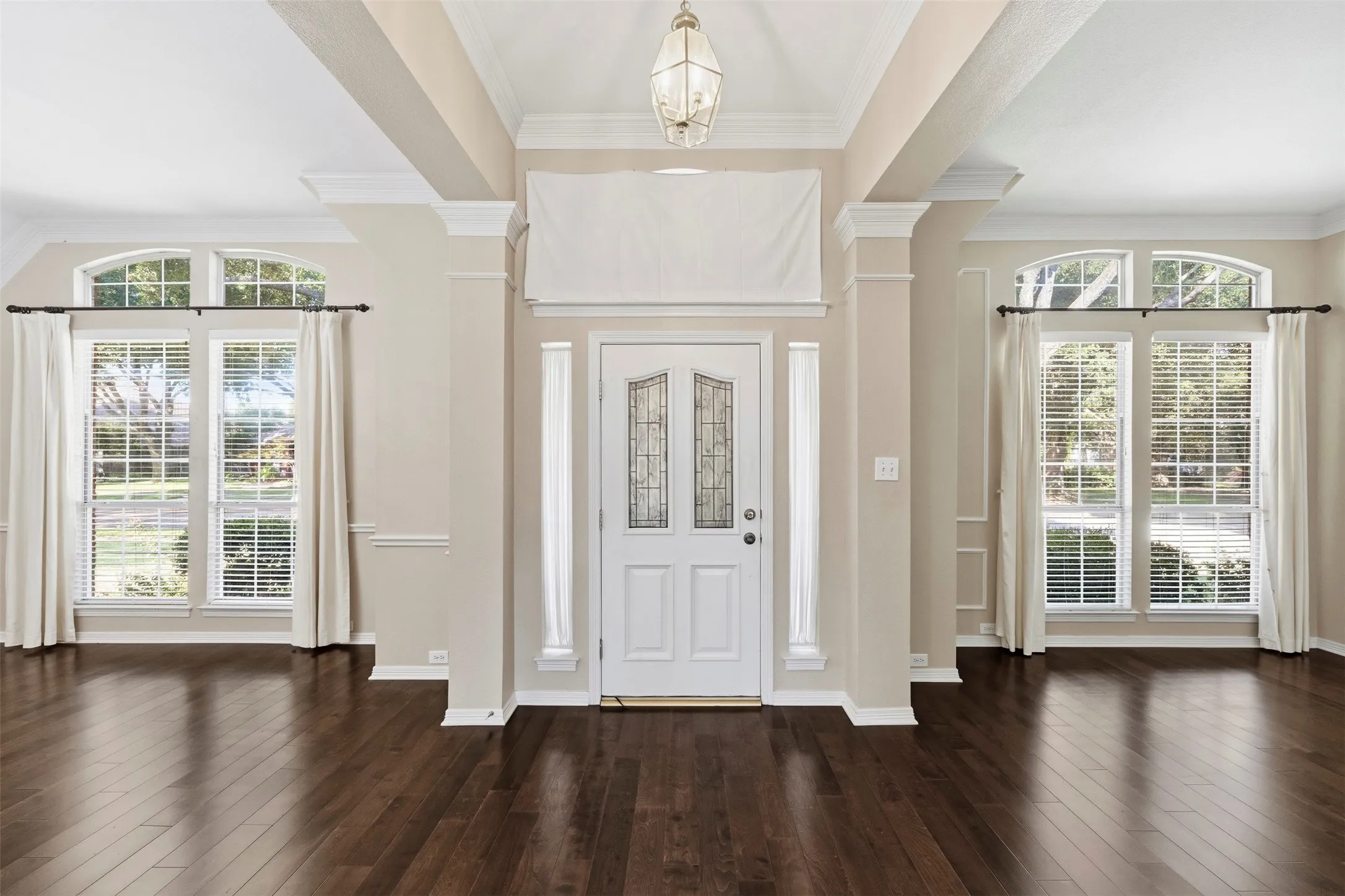 Dark wood-finish flooring, white paneled entry door with decorative glass, arched transom windows, crown molding, and a decorative pendant light fixture