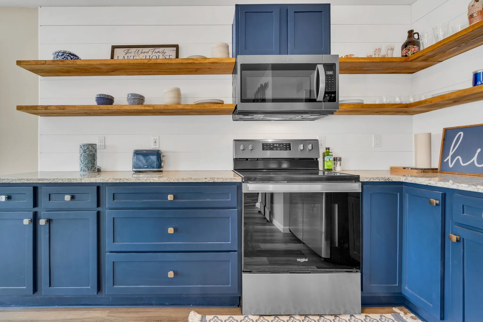 Blue cabinetry with square brass hardware, a granite-finish countertop, stainless steel appliances, and white shiplap-style backsplash