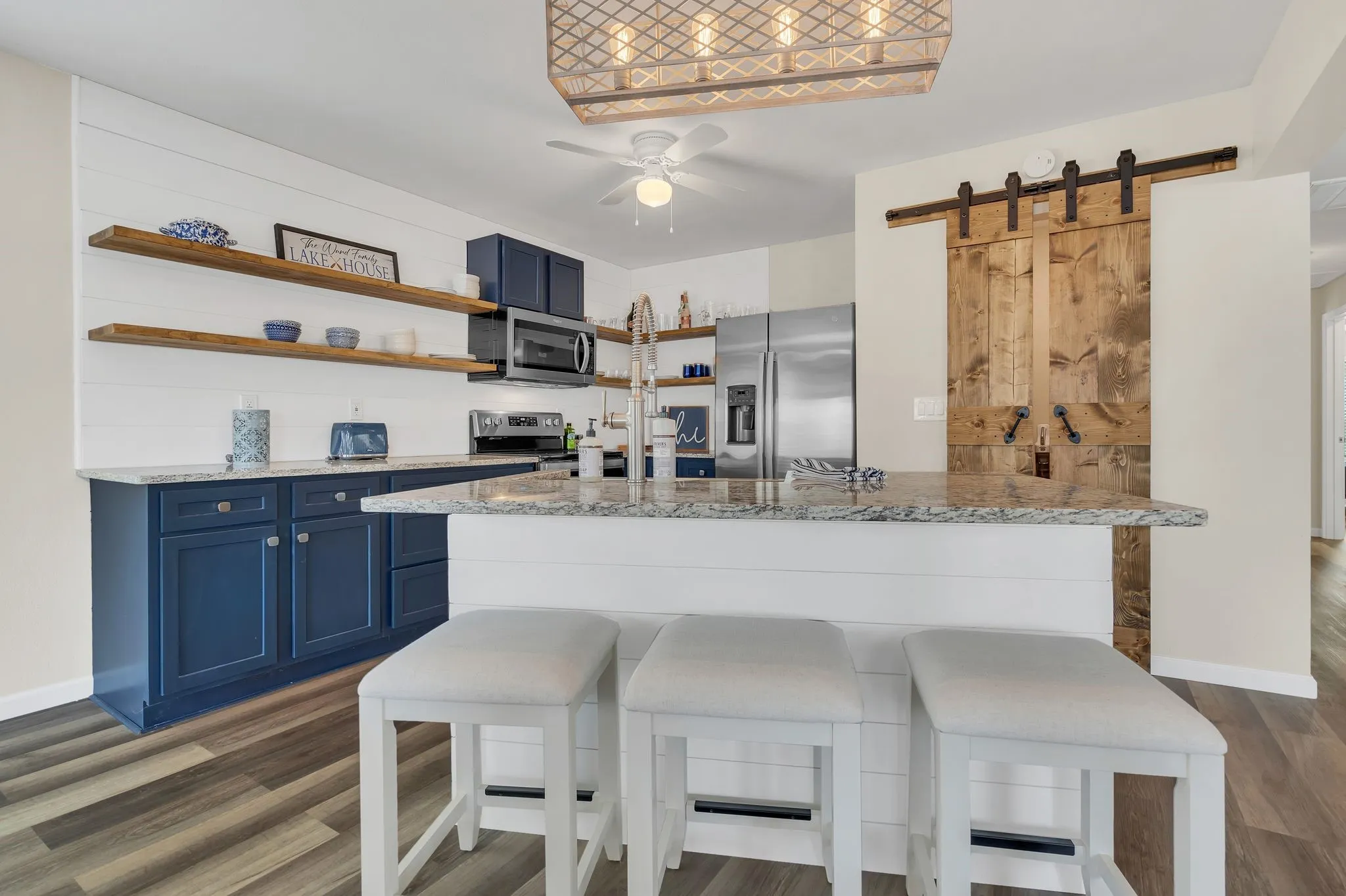 Expansive kitchen island with granite countertop, shaker-style blue cabinetry, stainless steel appliances, wood-finish flooring, and a sliding barn door
