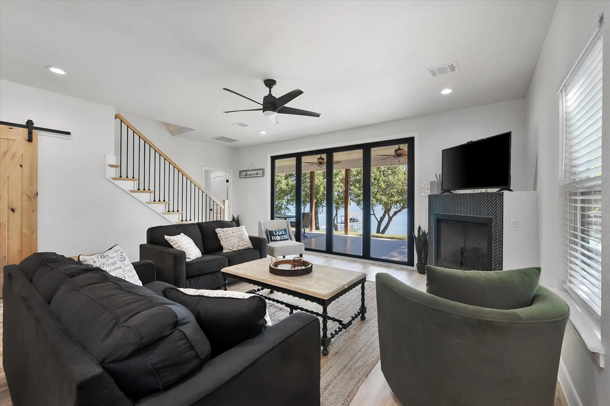 Spacious lake house living area combines comfortable gathering space, a fireplace, staircase, and wall of glass overlooking the waterfront.