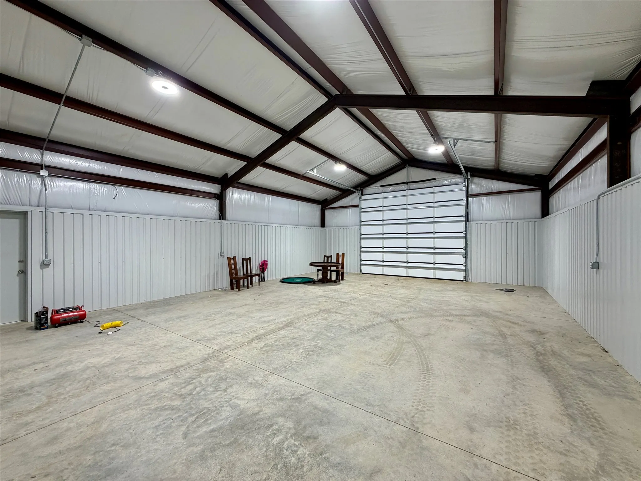 Spacious interior featuring a concrete floor, high vaulted ceilings with exposed steel beams, white corrugated metal walls, and an overhead garage door