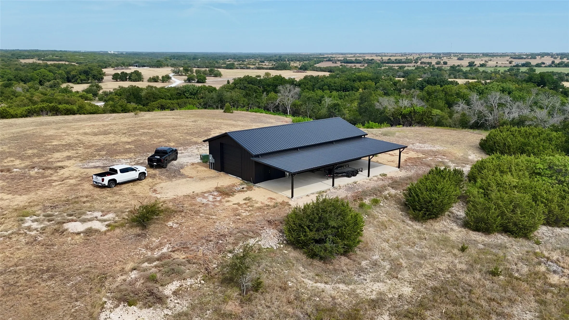 Black metal siding structure with a dark metal roof, featuring an attached covered concrete patio, surrounded by natural landscaping and expansive open land