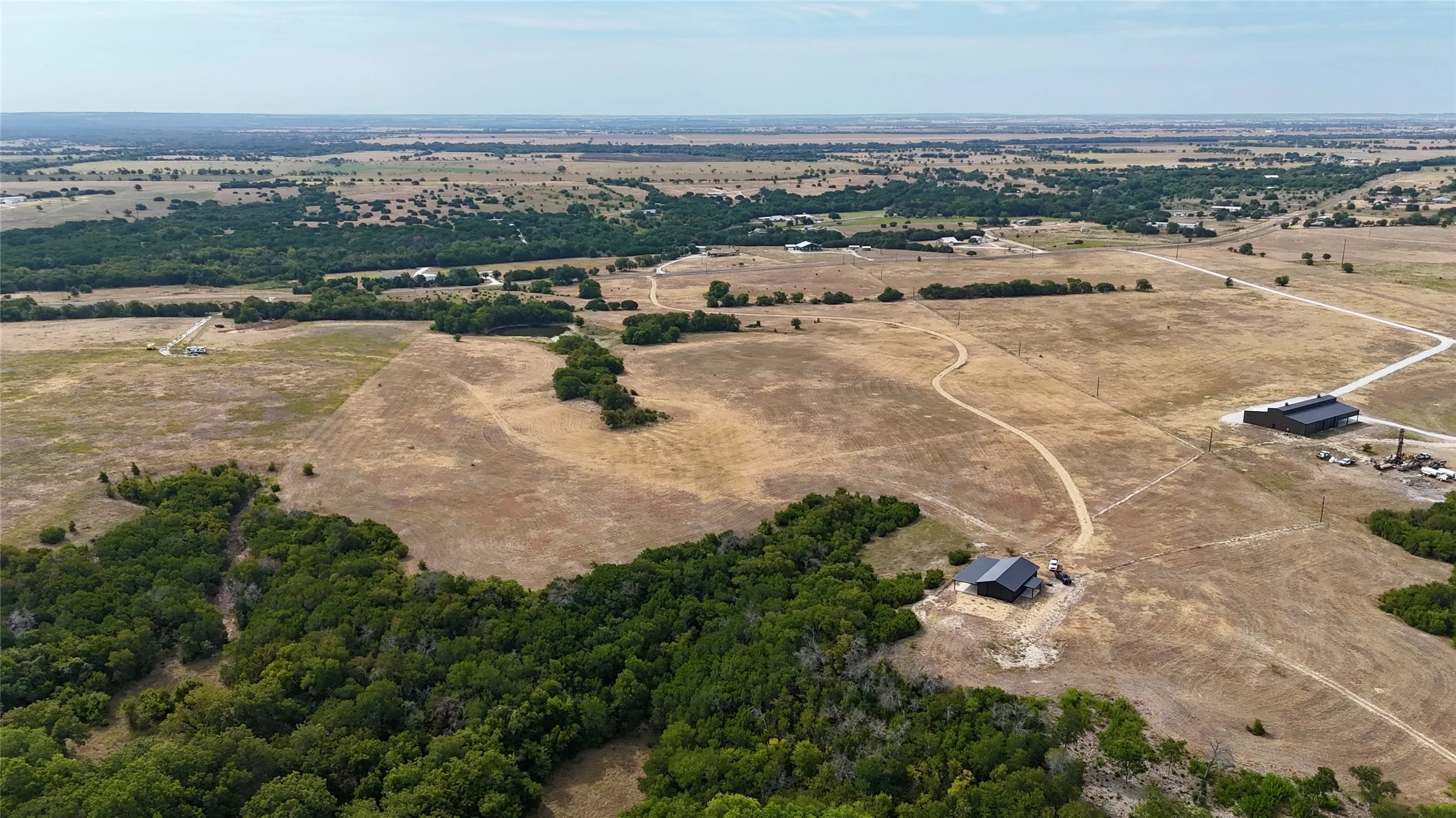 Expansive rural acreage featuring two structures with dark-toned roofs, surrounded by open fields and mature tree lines