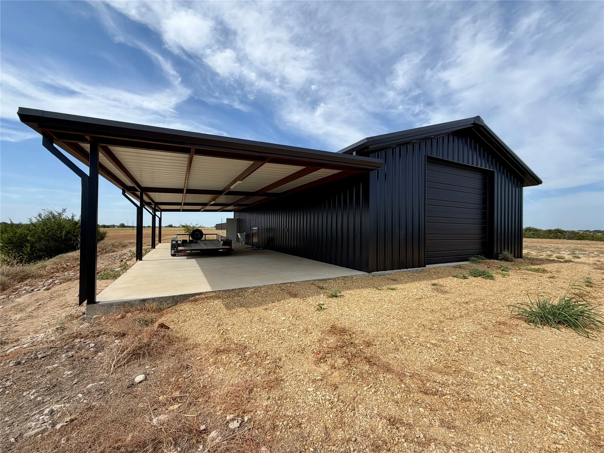 Modern black metal siding building featuring a large roll-up door, an attached covered concrete pad, and a pitched roof