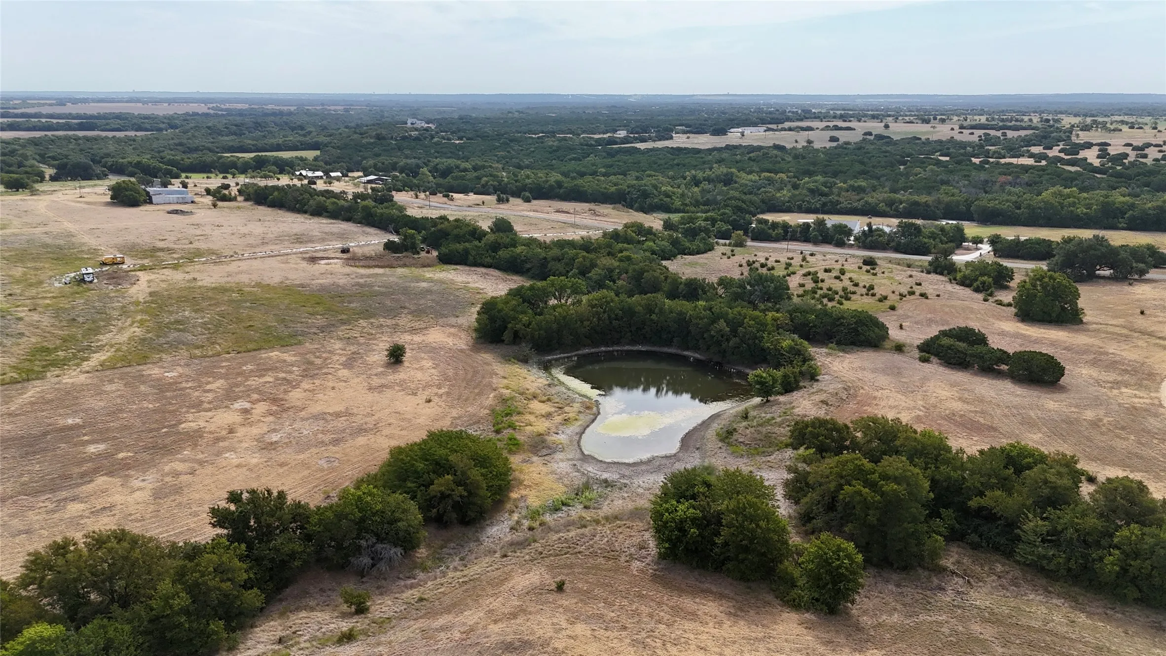 Expansive rural landscape featuring a pond, scattered trees, and distant structures