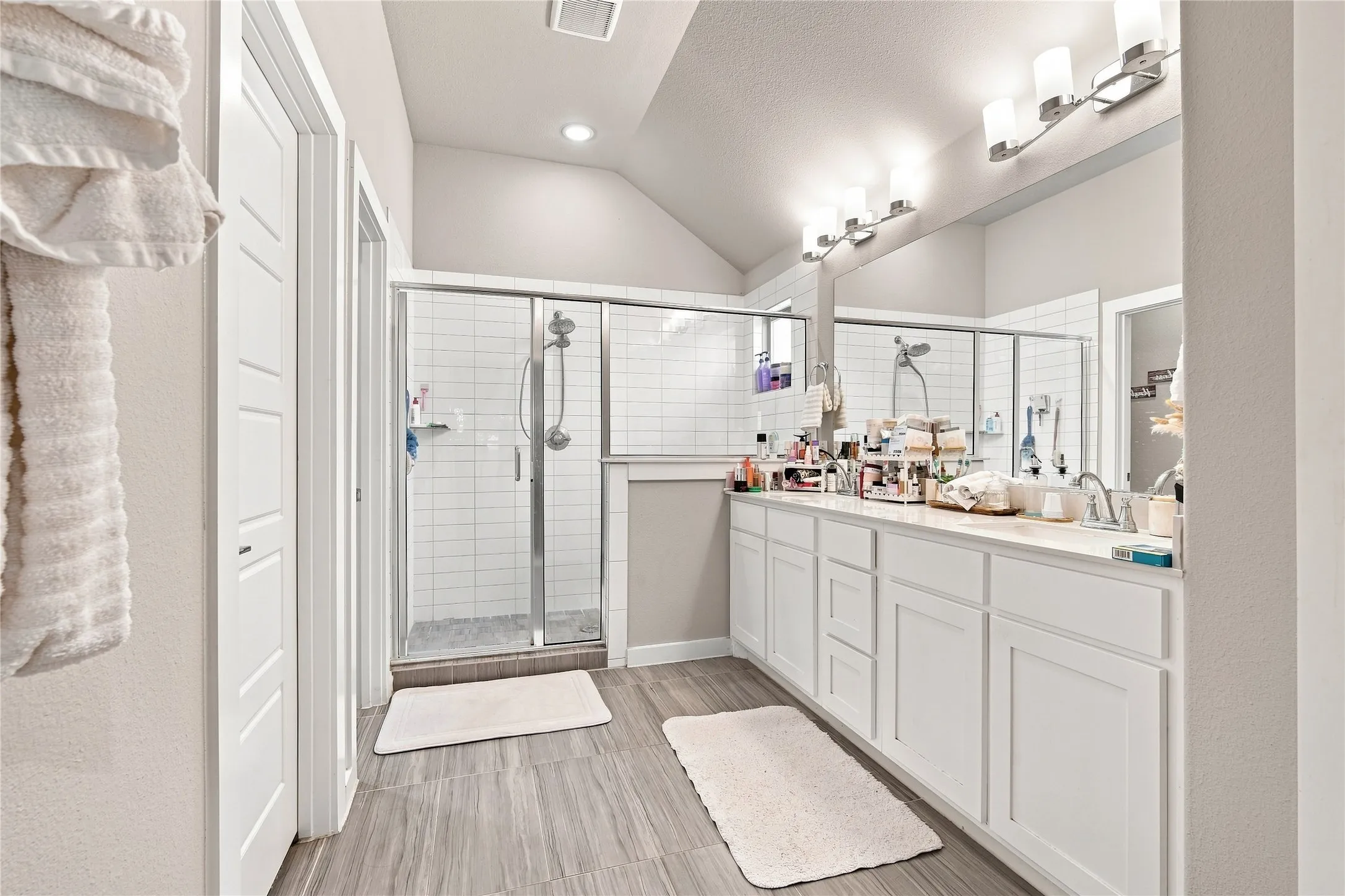 Spacious bathroom featuring a double vanity, large mirror, white cabinetry, a glass-enclosed shower with subway tile, and wood-finish flooring