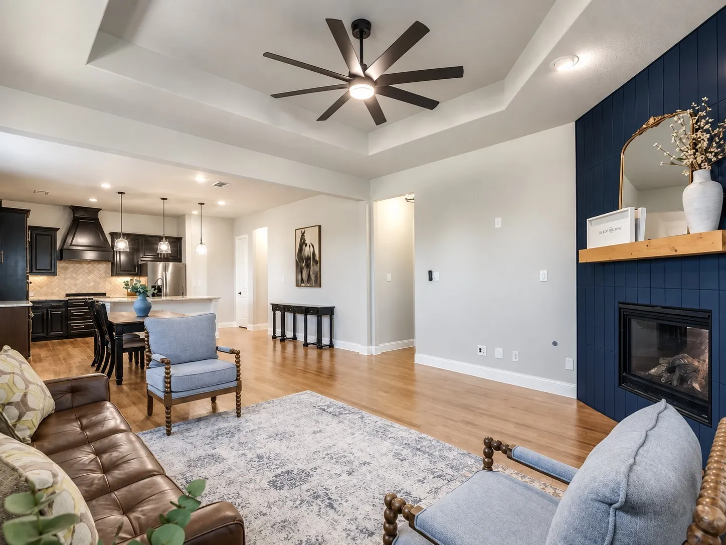 Tray ceiling with a ceiling fan, wood-finish flooring, fireplace with a blue tile surround, kitchen with dark cabinetry, and an island with pendant lighting