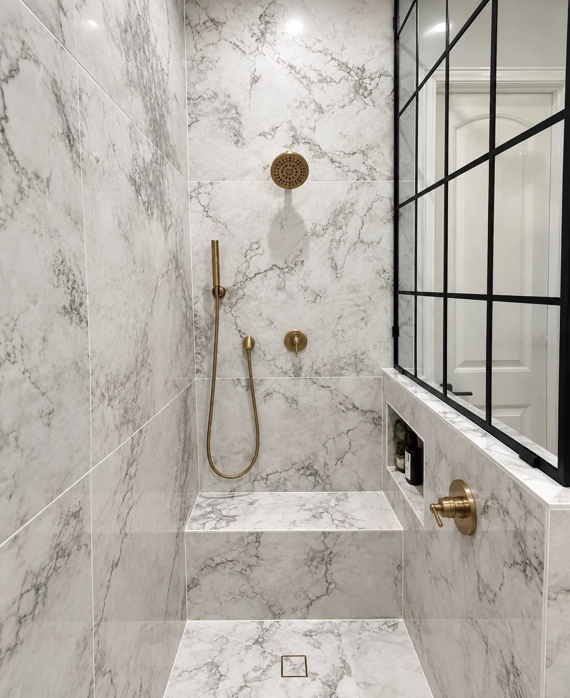 Shower featuring marble-look wall and floor tile, built-in bench, integrated niche, and brass fixtures