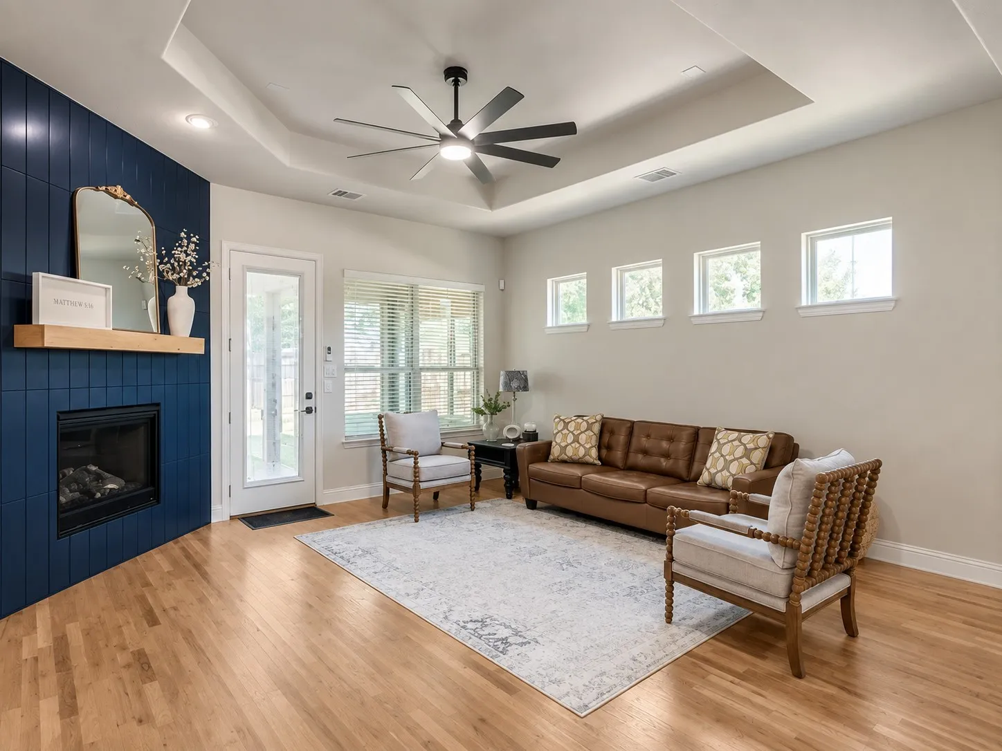 Hardwood flooring, a modern fireplace with a wood-finish mantel and blue tile surround, a tray ceiling, an integrated ceiling fan with lighting, and multiple windows