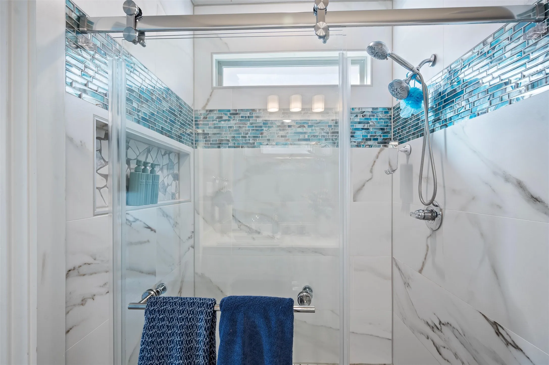 Shower enclosure featuring sliding glass doors, large format marble-look tile, a mosaic tile accent band, a built-in shower niche, and a fixed rainfall shower head with a handheld sprayer