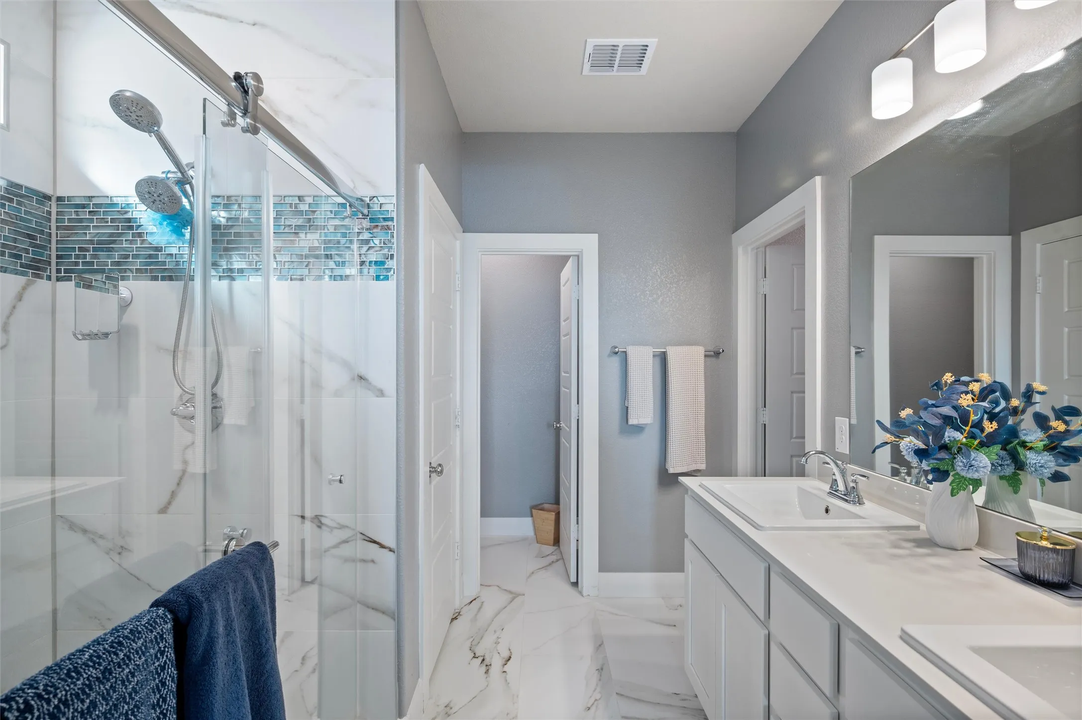Dual shower heads with a glass enclosure, white and blue tile accent, double vanity with integrated sinks, white cabinetry, and a large wall mirror