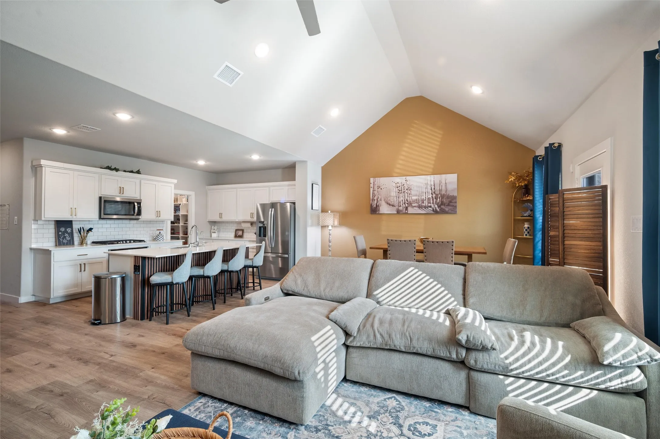Vaulted ceiling with recessed lighting, wood-finish flooring, white shaker cabinetry, stainless steel appliances, and a kitchen island with seating