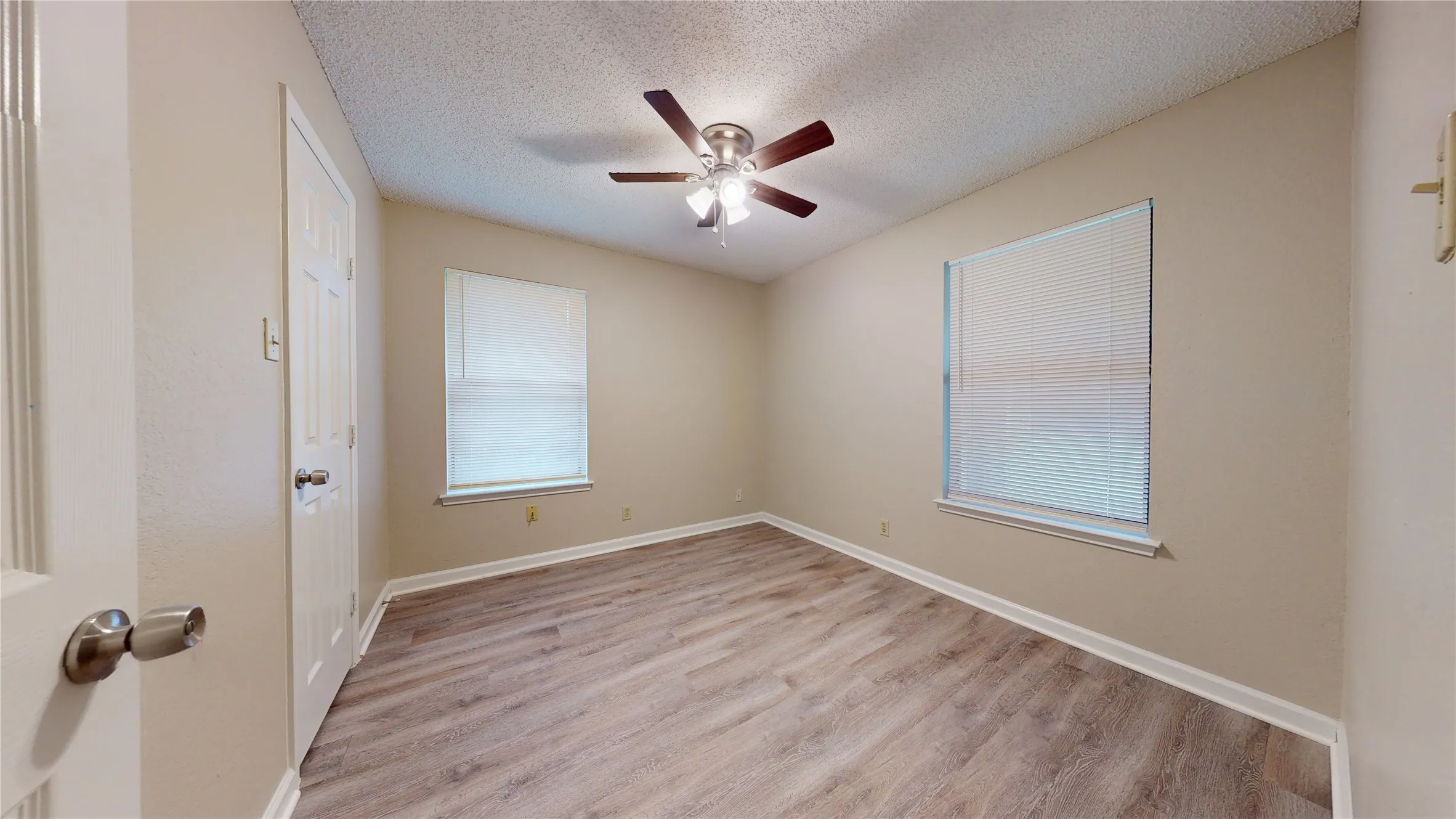 Room featuring wood-finish flooring, two windows with blinds, a ceiling fan, and a white interior door