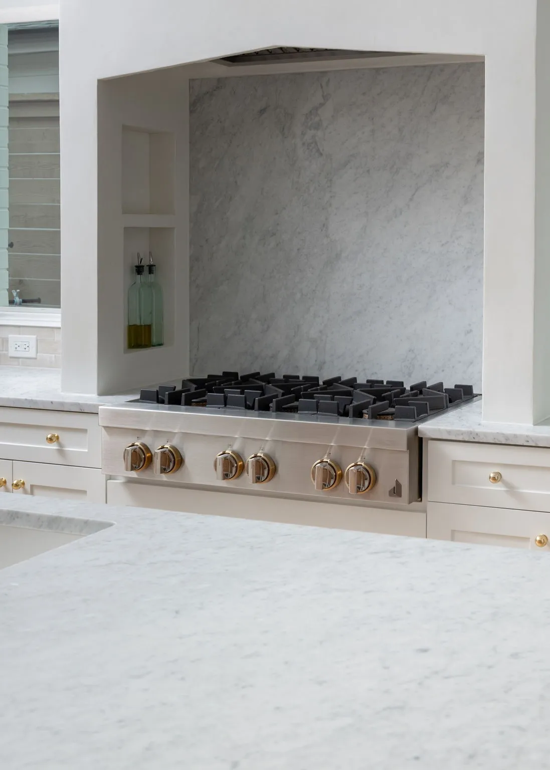 Built-in gas range with gold-tone hardware, integrated range hood, stone slab backsplash, stone countertops, and recessed shelving